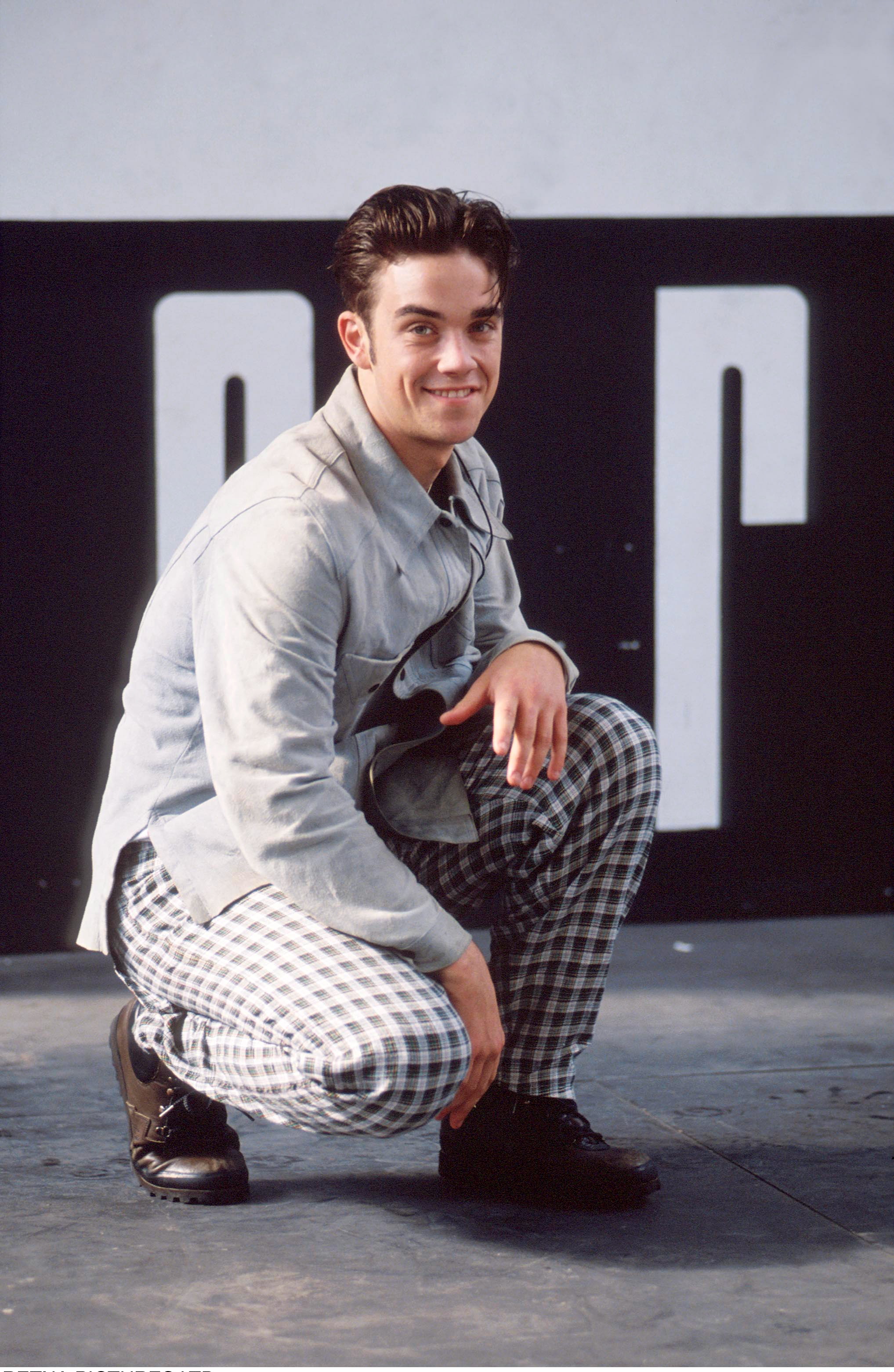 Robbie Williams s'accroupit pour un portrait en 1991, vêtu d'un pantalon à carreaux audacieux et d'une veste légère. Alors qu'il commence tout juste à s'imposer comme une idole des adolescents, son sourire espiègle et sa pose assurée reflètent l'arrogance précoce qui caractérisera ses premières années de gloire.