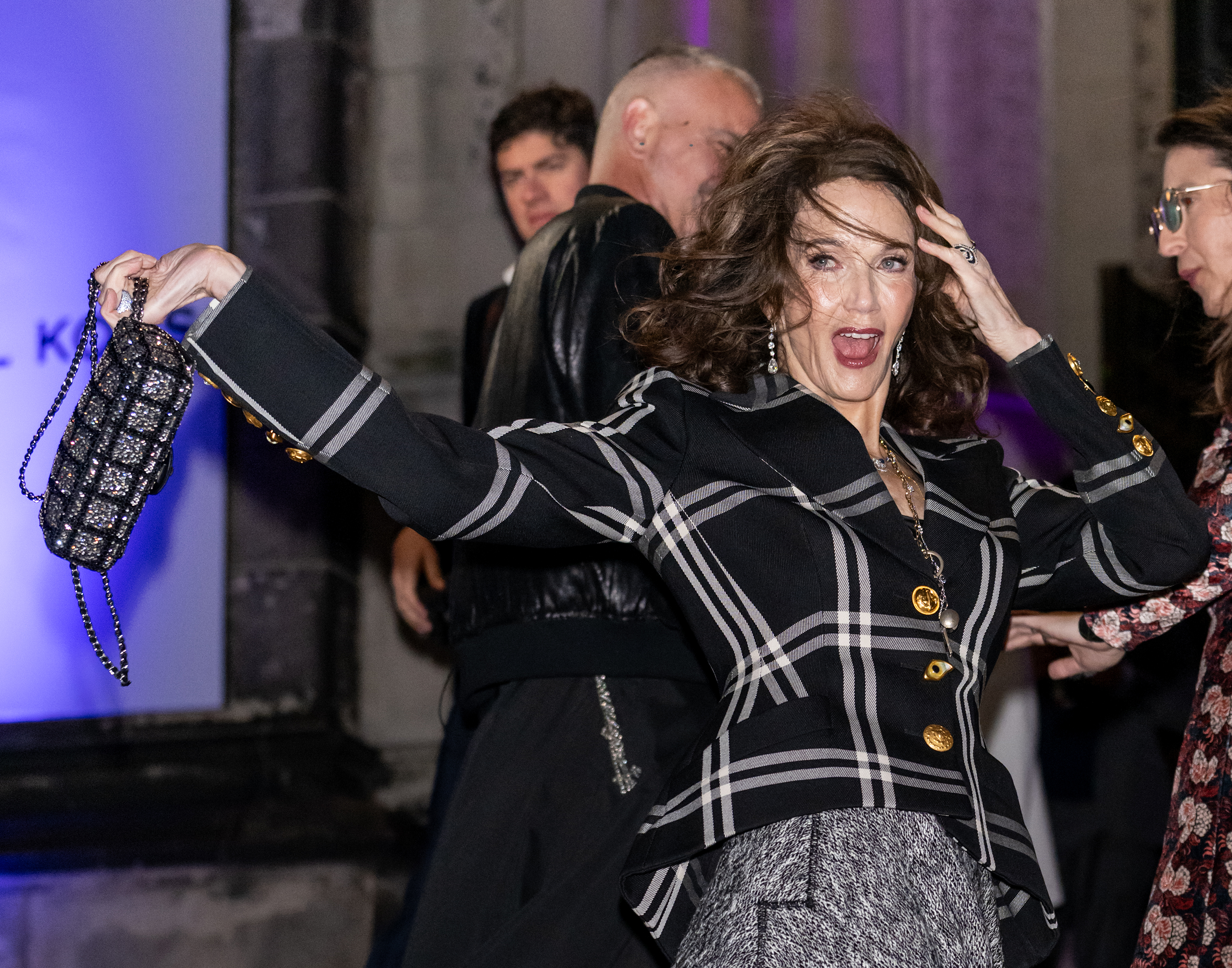 Lynda Carter prend une pose espiègle à l'extérieur de l'événement Michael Kors, secouant ses cheveux et exhibant un sac à main étincelant. Vêtue d'un blazer à carreaux audacieux avec des boutons dorés et d'une jupe grise texturée, elle dégage une énergie et un charisme incroyables tout en ravissant les photographes avec son style animé.