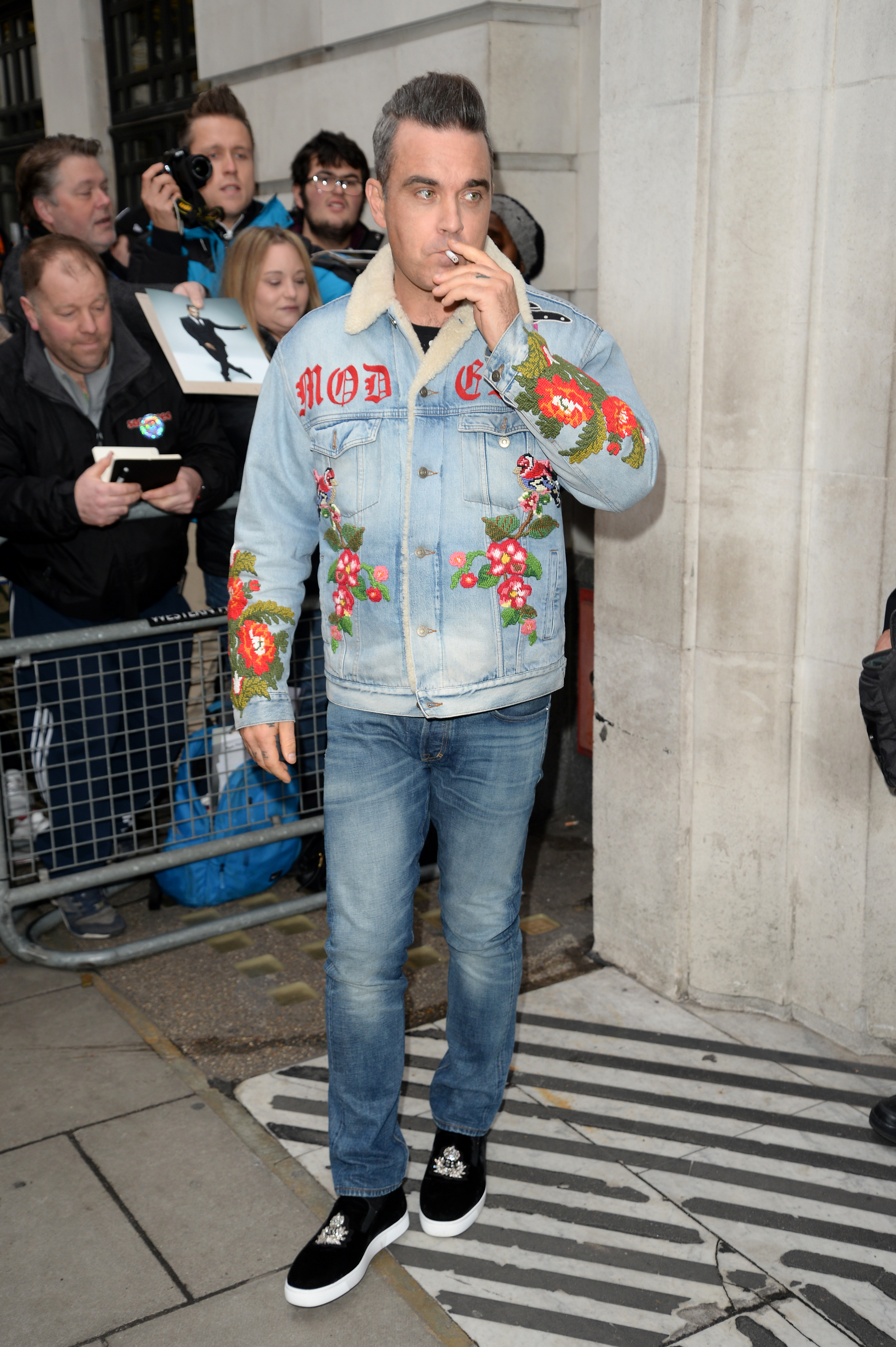 Robbie Williams aperçu devant les locaux de BBC Radio 2 le 8 décembre 2017 à Londres, en Angleterre. Vêtu d'une veste en jean à fleurs et fumant une cigarette, il a affiché un style audacieux tout en saluant les fans rassemblés devant le studio.