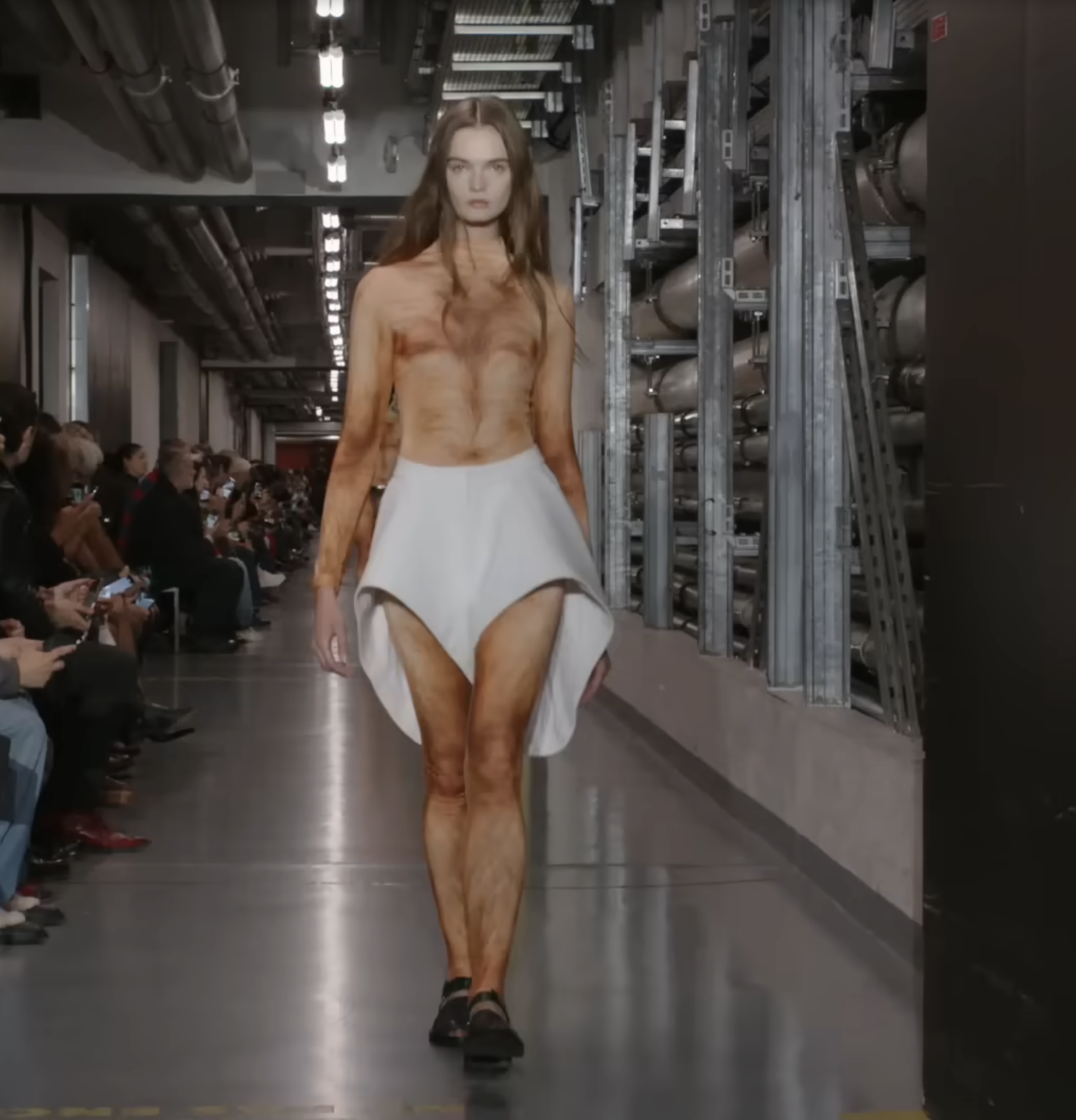 Un autre mannequin masculin a fait une entrée remarquée dans une jupe blanche asymétrique, plus longue à l'arrière qu'à l'avant. Il semble s'agir de son seul vêtement, car il défile avec assurance sur le podium. Extrait d'une publication datée du 5 octobre 2025. Source : Youtube/@jeanpaulgaultier