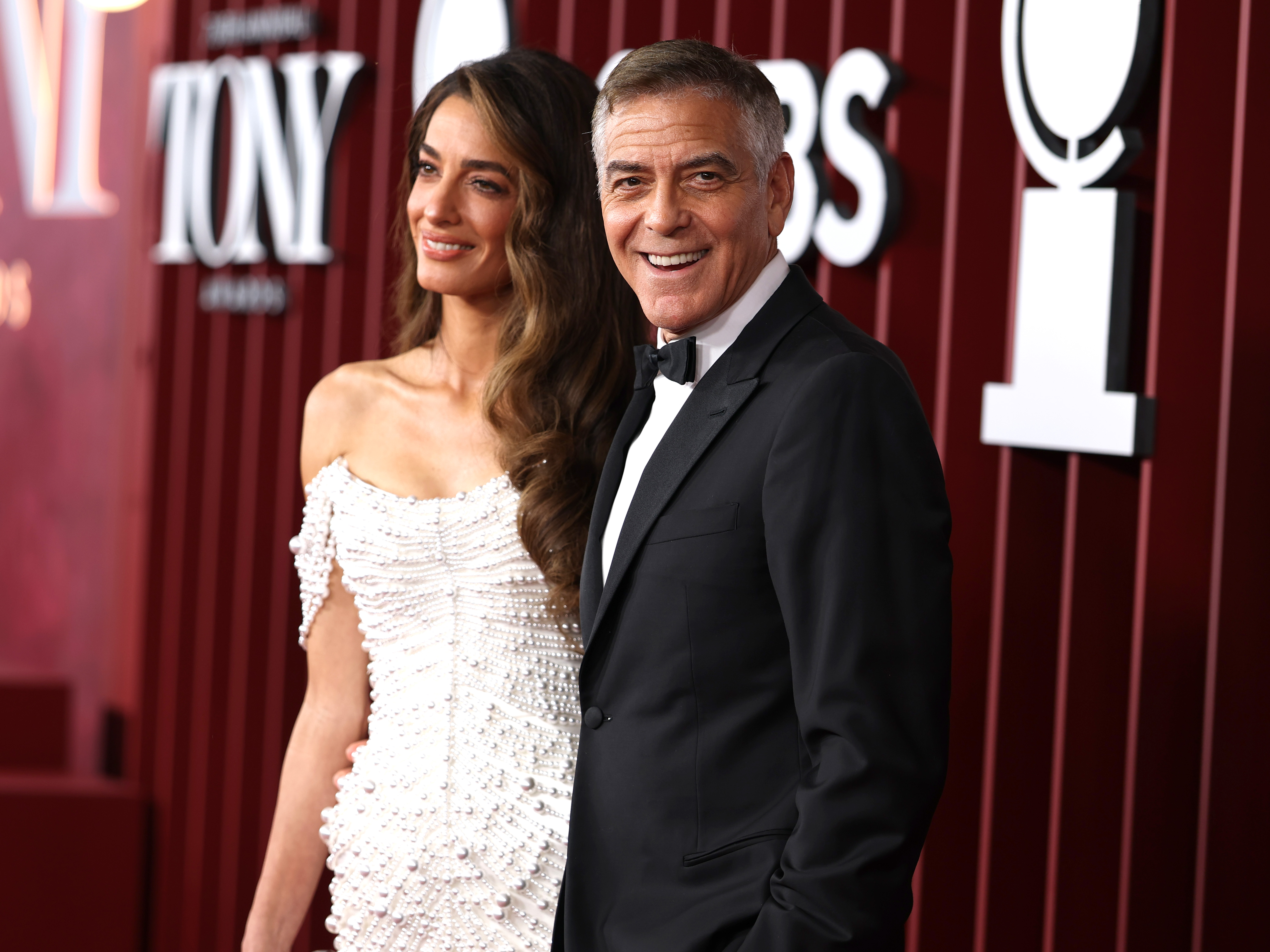 George Clooney sourit doucement aux caméras tandis qu'Amal Clooney se tient à ses côtés. Sa robe drapée de perles brille sous les lumières alors qu'ils posent ensemble sur le tapis rouge des Tony Awards.