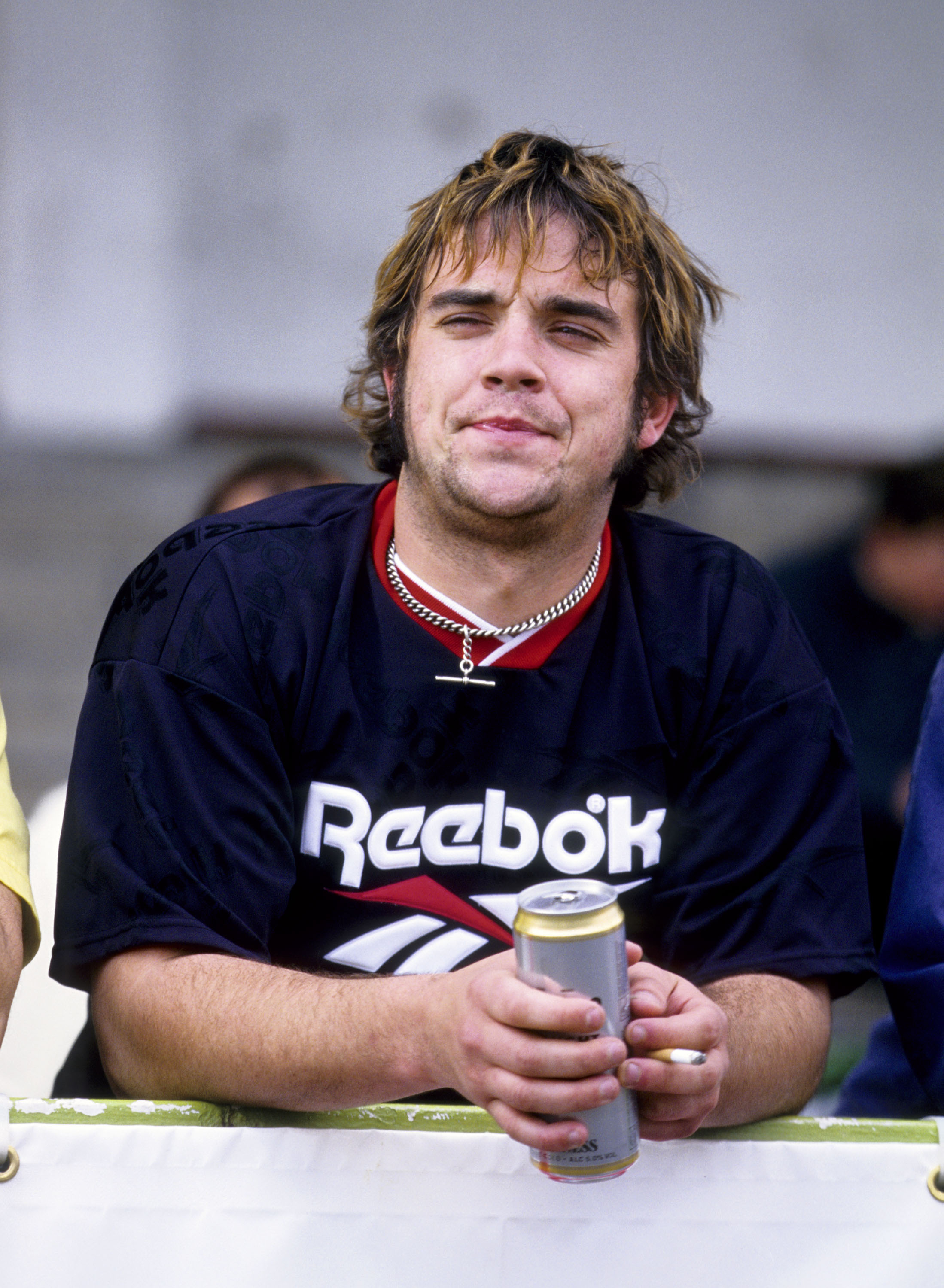 Robbie Williams assiste à un match de football caritatif en mai 1996, une bière et une cigarette à la main, vêtu d'un maillot Reebok. Ce moment de franchise reflète l'image rebelle qu'il s'est forgée après avoir quitté Take That.