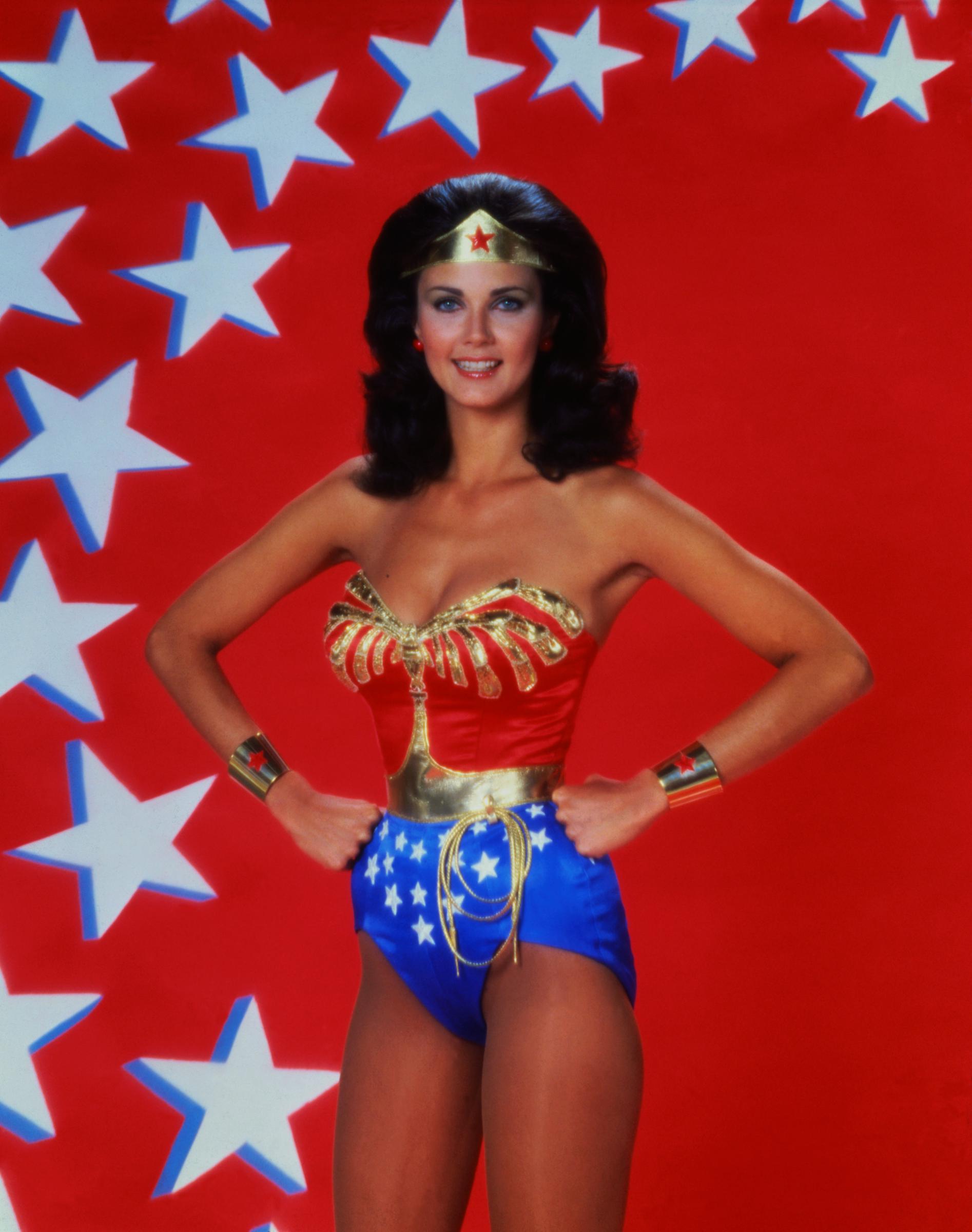 Lynda Carter se tient fièrement dans son costume classique de « Wonder Woman » devant un fond rouge vif parsemé d'étoiles blanches. Les mains sur les hanches, elle arbore un sourire éclatant tout en portant la version modernisée de la tenue emblématique de la super-héroïne, composée d'un corsage doré orné d'un aigle, d'un short bleu parsemé d'étoiles et de sa tiare caractéristique.