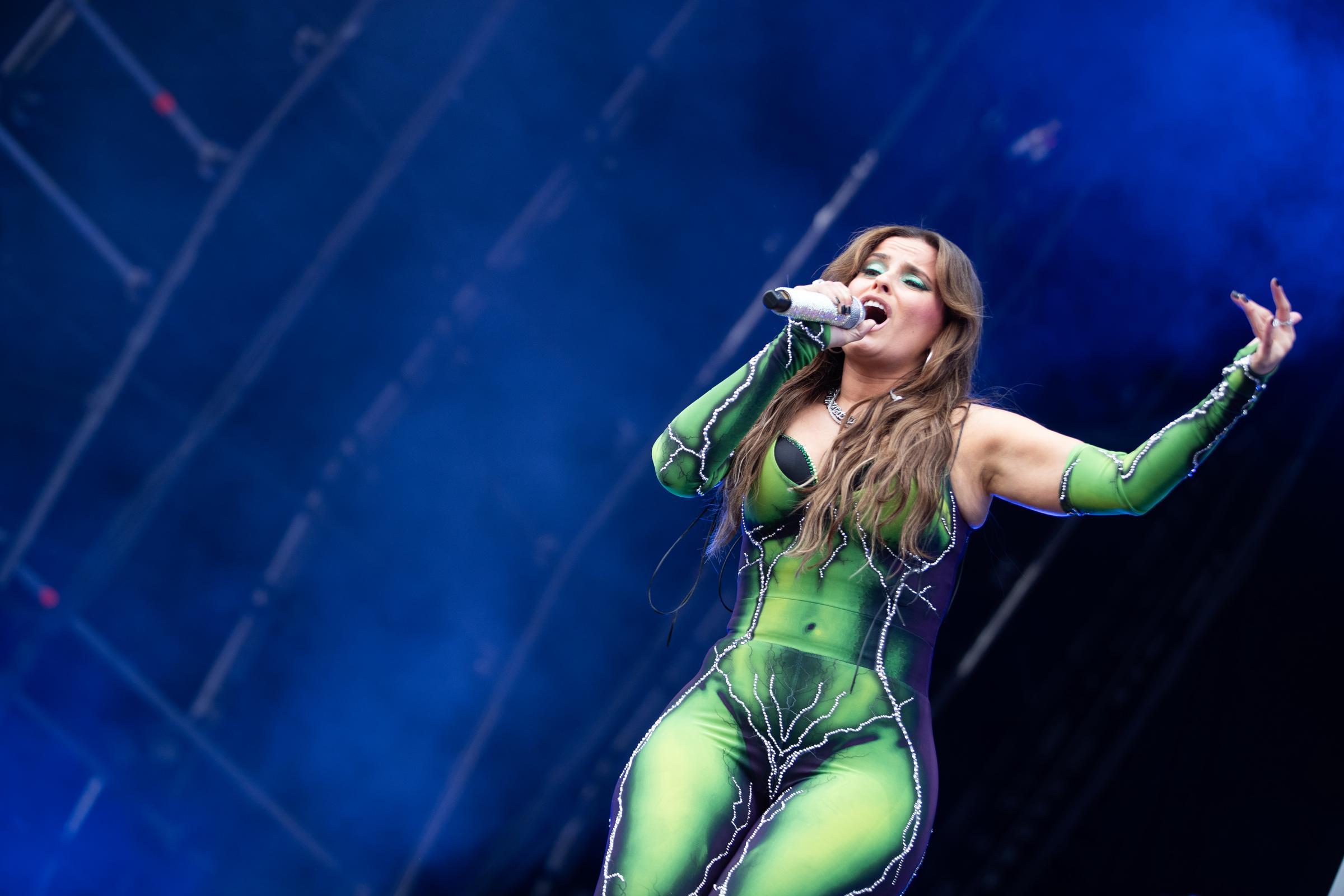 Furtado se produit dans une combinaison verte et noire ornée de détails argentés et de découpes. Elle tient un micro dans une main tout en chantant, avec un éclairage bleu derrière elle.