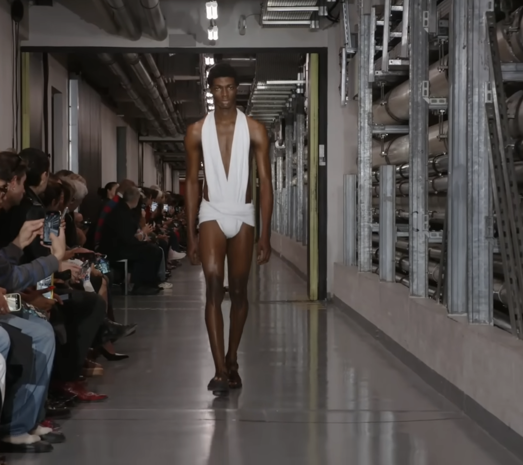 Un mannequin masculin vêtu d'un body ajouré. Cette fois-ci, il est blanc, contrastant de manière saisissante avec sa peau couleur chocolat noir. Le look suggère que la mode futuriste pourrait s'inspirer du passé, avec une partie au niveau des reins qui rappelle le subligaculum romain. Extrait d'un post daté du 5 octobre 2025. Source : Youtube/@jeanpaulgaultier