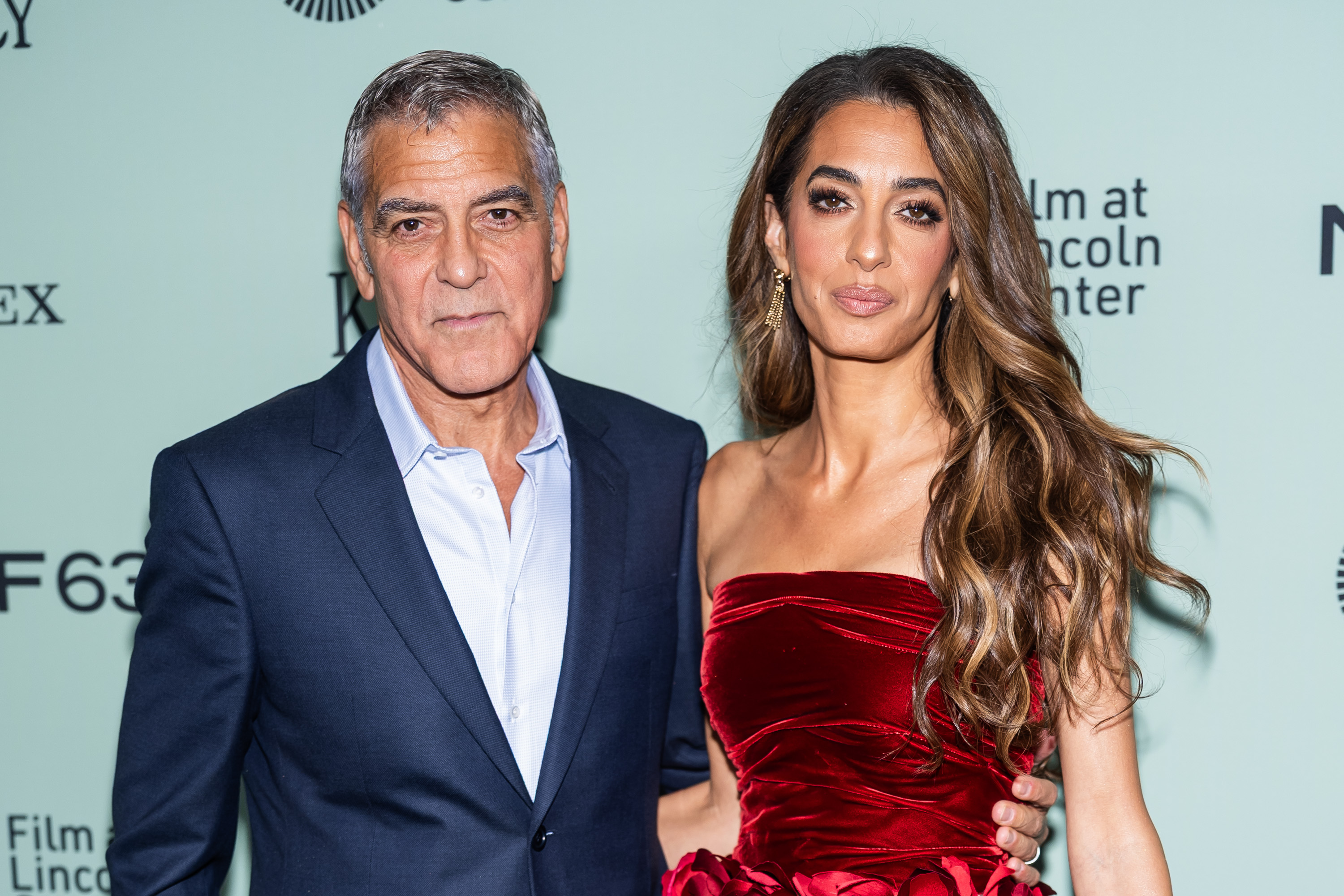 George Clooney et Amal Clooney font face côte à côte au mur de journalistes. Elle affiche un sourire discret et serein, tandis qu'il la regarde avec une expression calme, mais indéniablement fière.