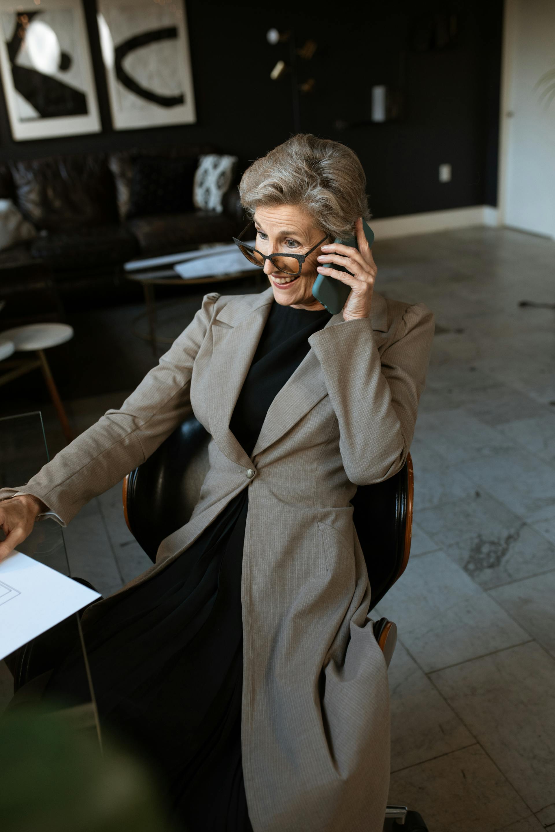 Une femme d'âge mûr souriante, vêtue d'un manteau bronzé et portant des lunettes, assise sur une chaise et parlant au téléphone | Source : Pexels
