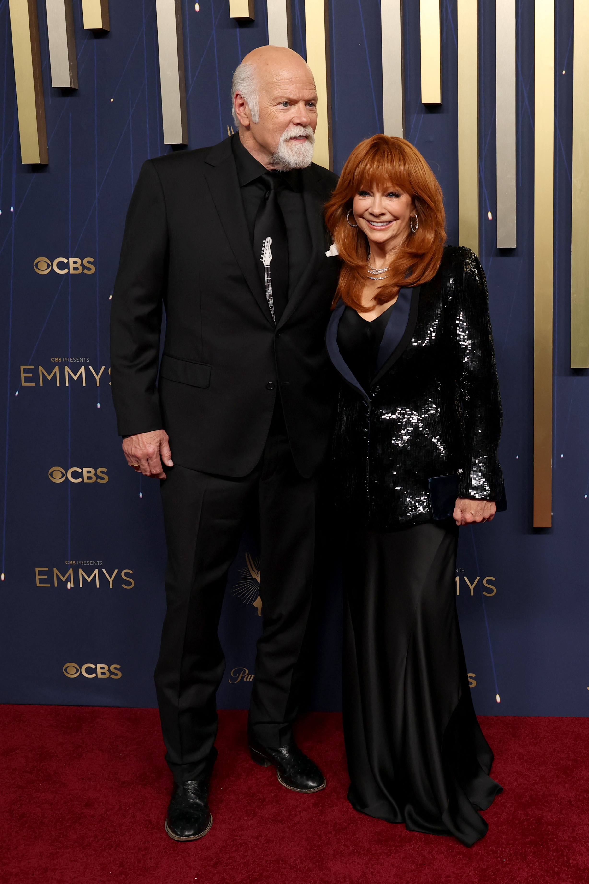 Reba McEntire, vêtue d'un blazer noir à paillettes sur une longue robe en soie noire, pose main dans la main avec Rex Linn aux Emmys. Linn a complété son look avec un costume noir, une cravate brodée de blanc et un revers blanc.