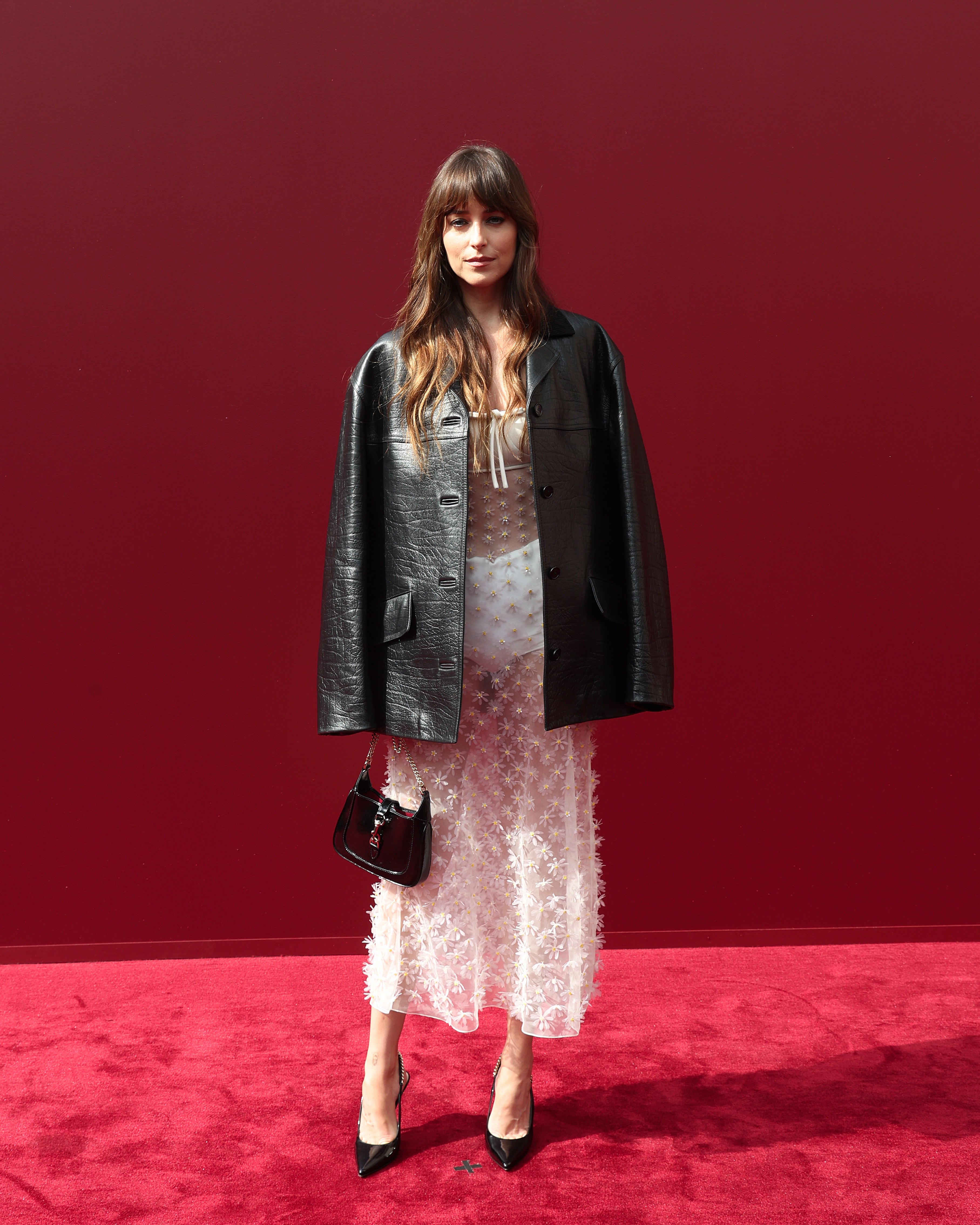 Dakota Johnson assiste au défilé printemps-été de Gucci Women, resplendissante dans une robe blanche transparente à fleurs portée sur une combinaison. Associée à une veste en cuir noir oversize, un sac à main verni et un maquillage discret, elle affiche un look à la fois moderne et vintage.
