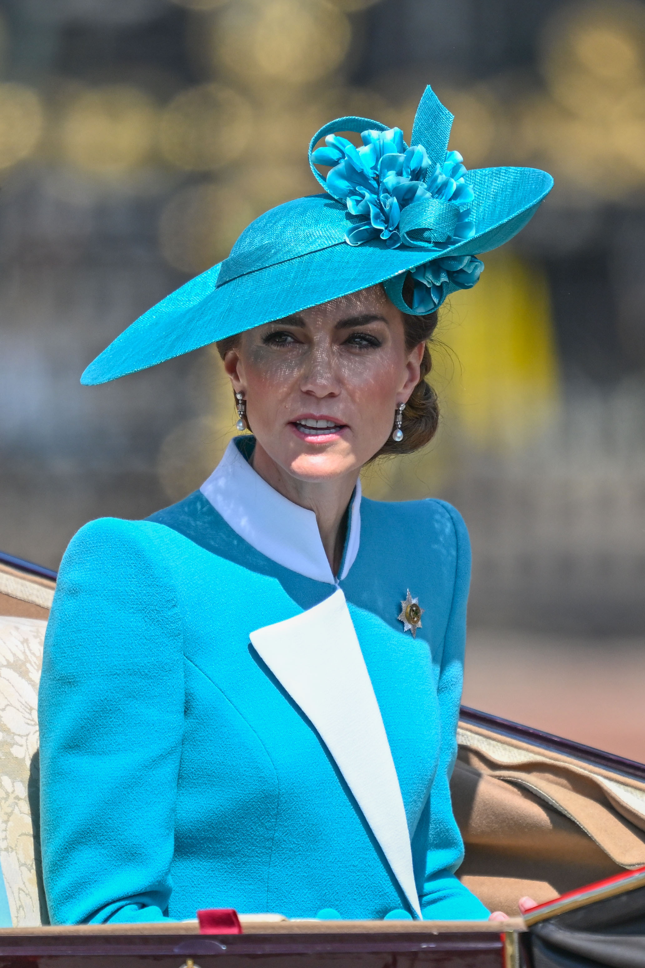 Dans ce plan moyen, on voit la princesse Catherine vêtue d'une tenue bleu sarcelle et blanche lors de la parade Trooping the Colour de 2025. Le design structuré de son blazer est clairement visible, avec des accents blancs audacieux au niveau du col et des poignets qui contrastent avec le tissu bleu sarcelle riche. Posée et sereine, elle incarne l'élégance royale moderne.