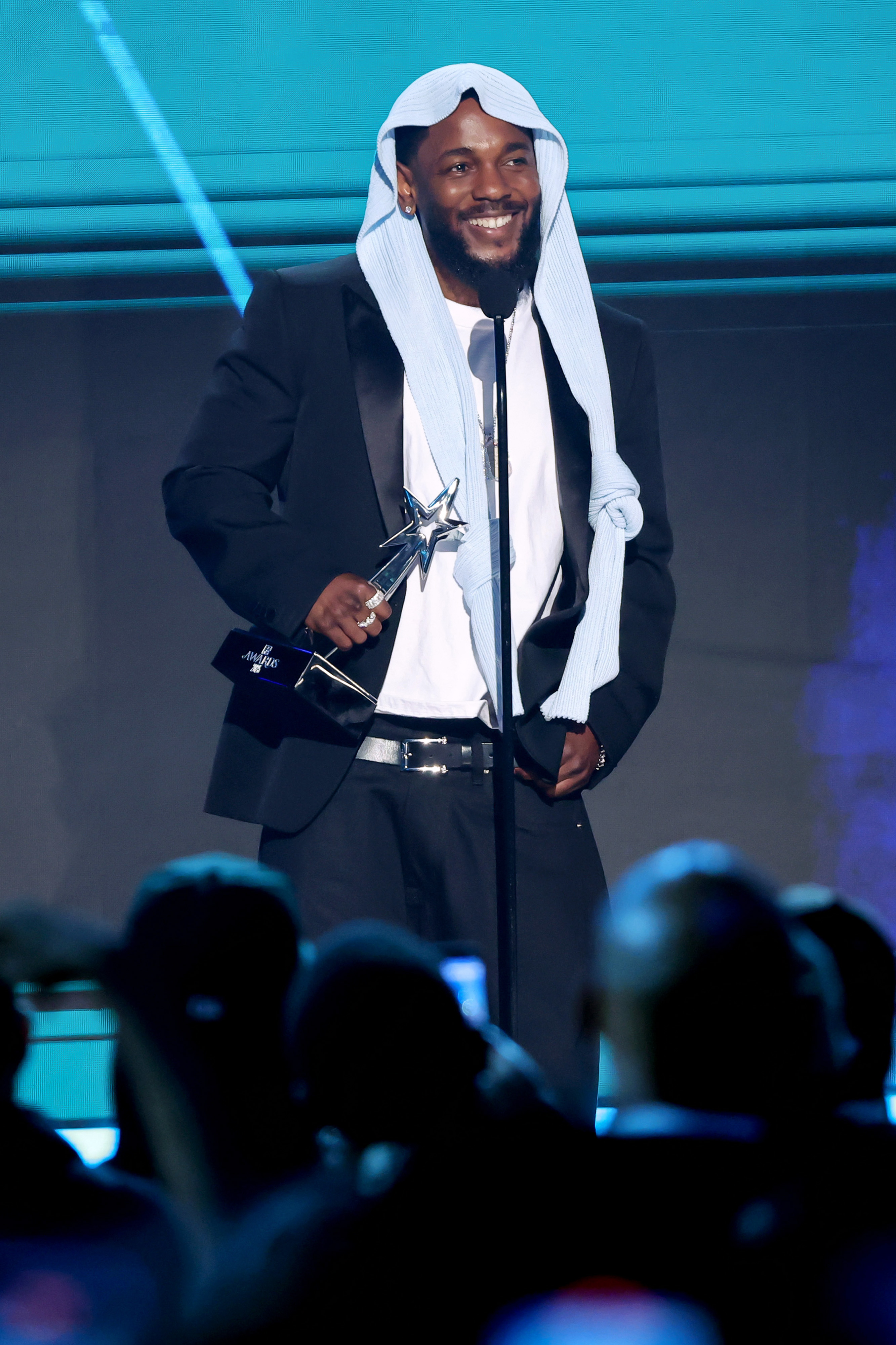 Kendrick Lamar affiche un sourire éclatant alors qu'il tient un trophée sur la scène des BET Awards 2025. Vêtu de son couvre-chef désormais iconique et de son costume noir, le rappeur semble véritablement ému par la reconnaissance continue de la soirée.