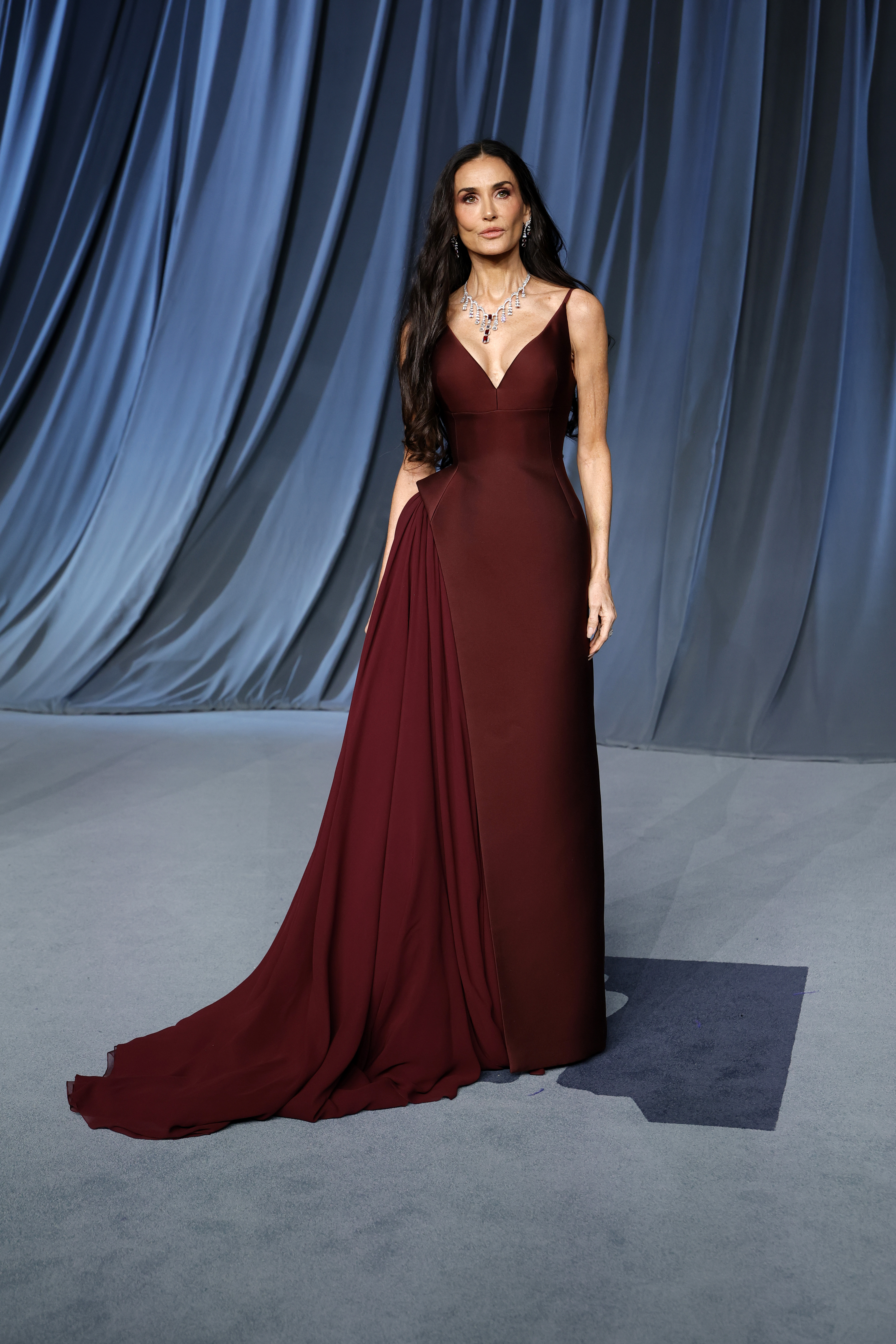 Demi Moore se tient droite, le regard posé devant elle, les bras légèrement le long du corps, tandis que sa somptueuse robe bordeaux tombe en cascade autour de ses pieds. La coupe impeccable de la robe met pleinement en valeur son arrivée sur le tapis rouge.