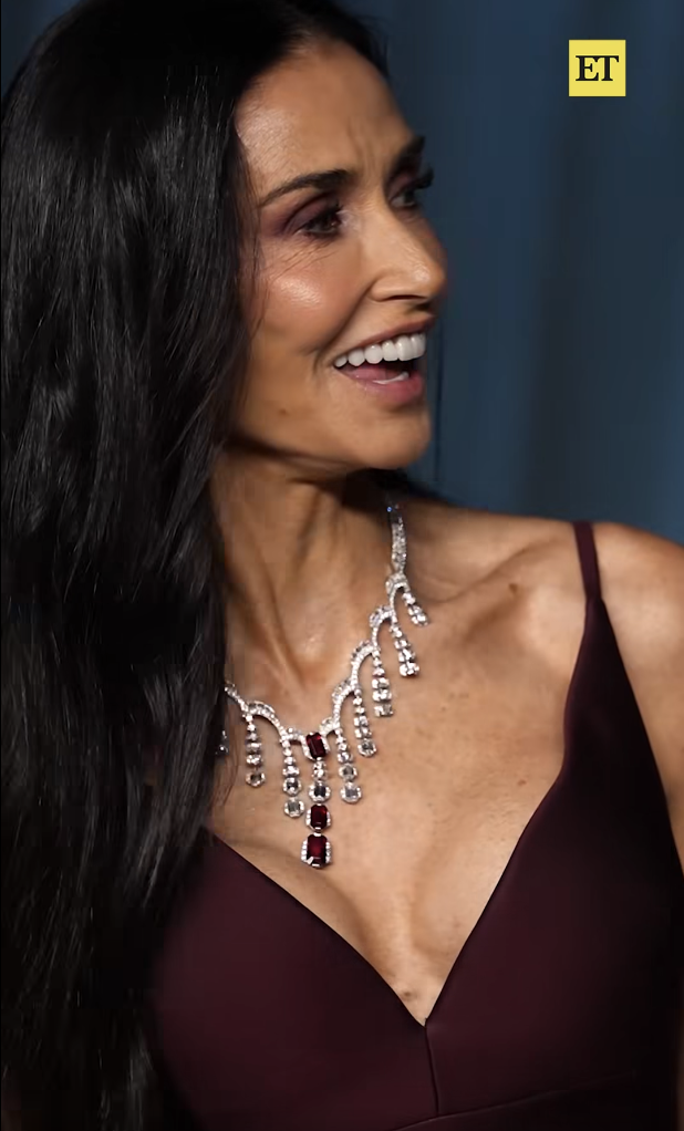 Surprise en plein rire, Demi Moore se tourne vers sa droite dans un moment de spontanéité, le regard plein de chaleur, tandis que la lumière se reflète sur son collier imposant et les riches nuances de sa robe bordeaux. | Source : Facebook/EntertainmentTonight