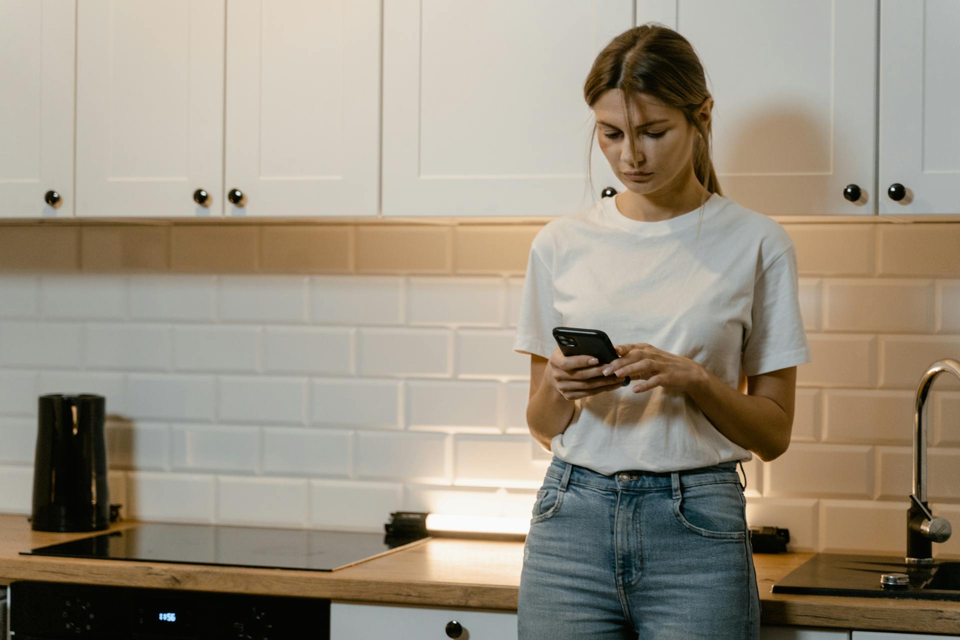 Une femme vêtue d'un t-shirt blanc et d'un jean utilise un téléphone alors qu'elle se tient debout dans une cuisine | Source : Pexels