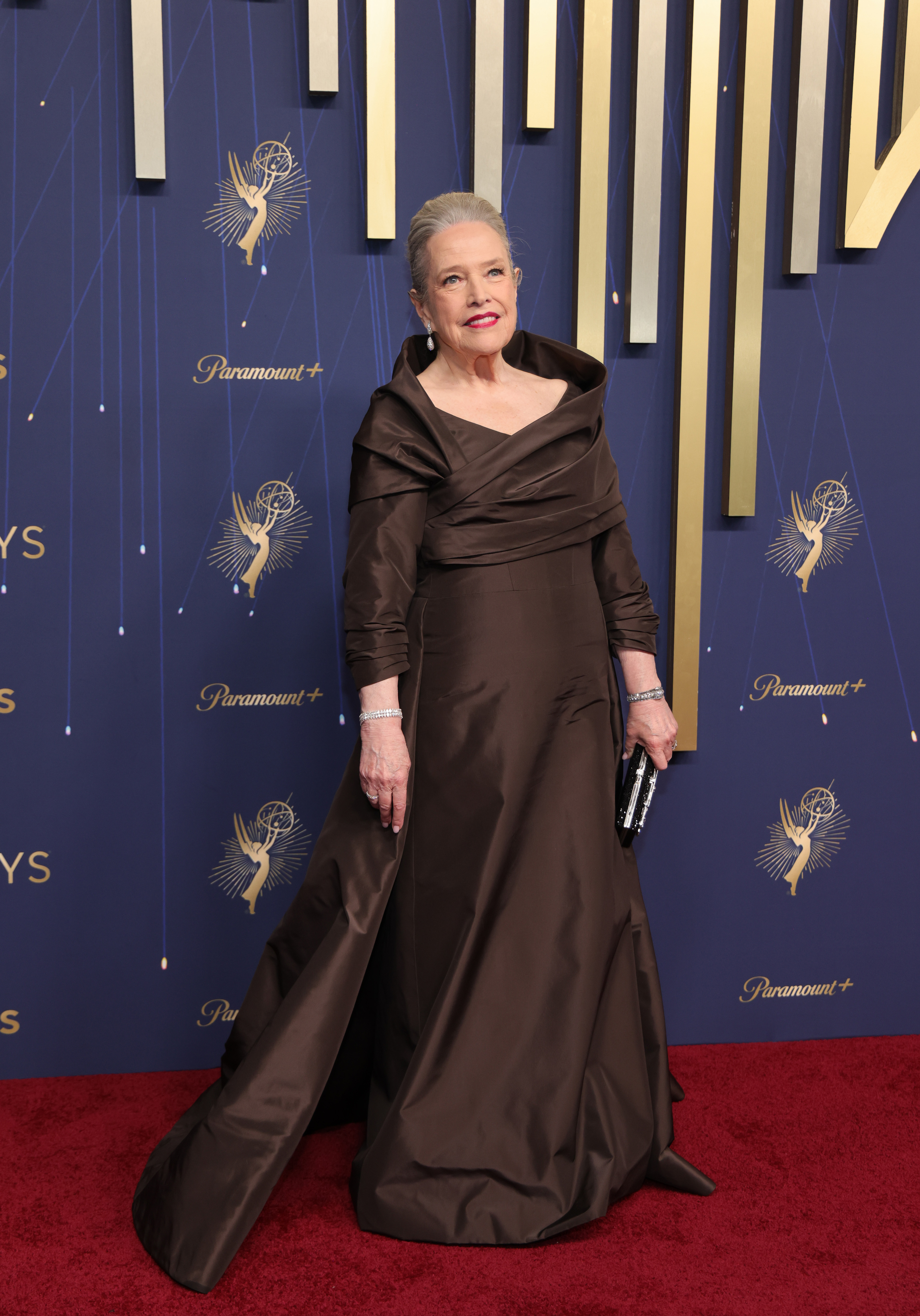 Kathy Bates portait une robe longue raffinée de couleur sombre avec un décolleté ouvert aux Emmys. Elle a accessoirisé sa tenue avec un chignon volumineux, un rouge à lèvres rouge vif et des bijoux en or blanc et diamants, notamment un double bracelet tennis avec des pierres taillées en poire.