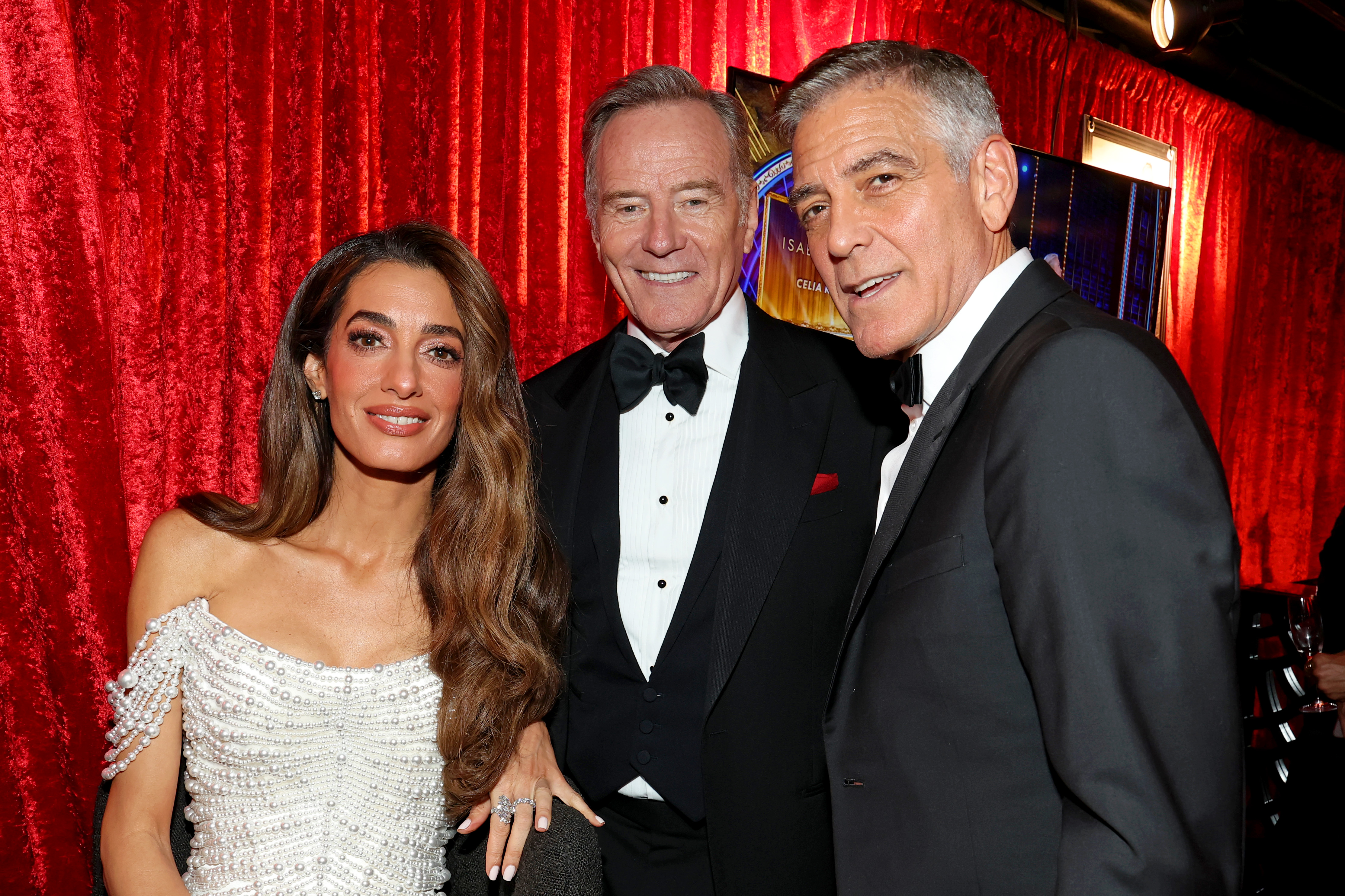 Amal et George Clooney rejoignent Bryan Cranston dans les coulisses de la salle verte. Encadrée par des rideaux de velours rouge, Amal sourit directement à la caméra, tandis que George se penche vers elle avec une expression détendue.