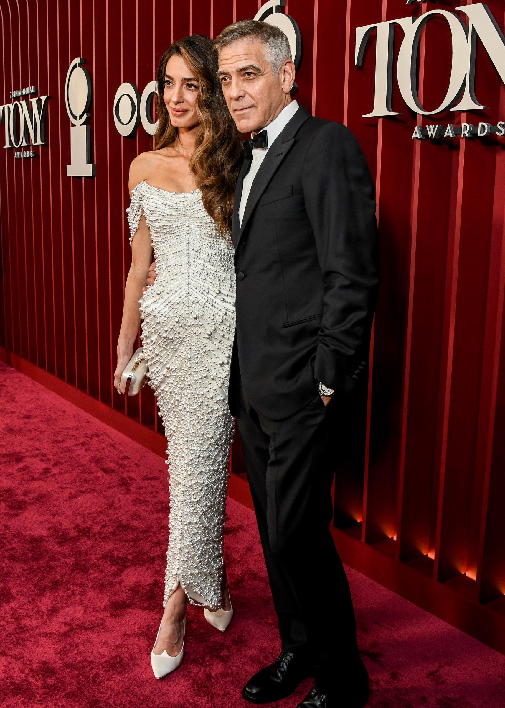 Amal Clooney se tient debout, les pieds croisés, à côté de George Clooney lors de la cérémonie des Tony Awards. George semble détendu alors qu'ils posent ensemble devant la toile de fond d'un rouge profond.