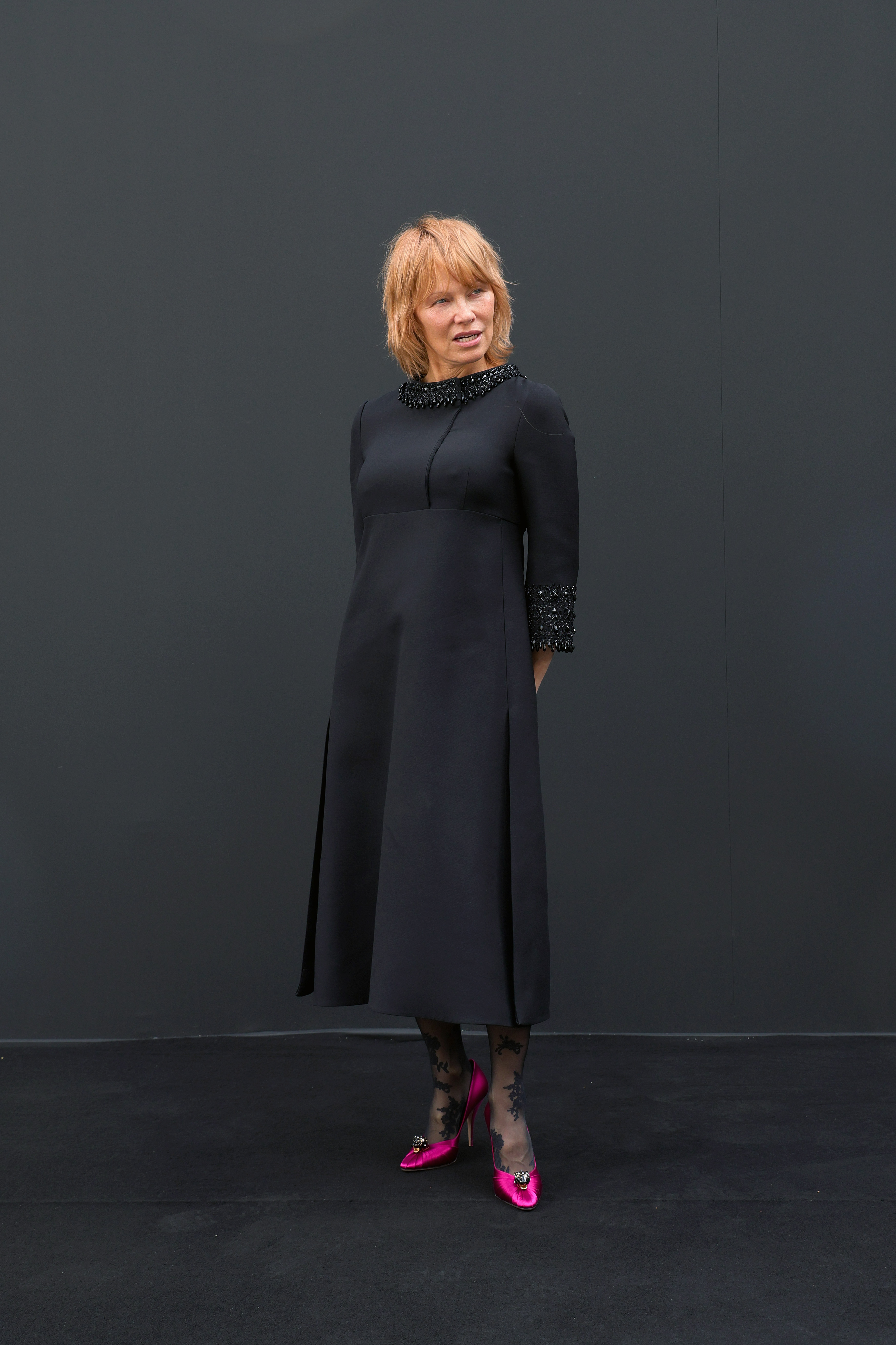 Pamela Anderson assiste au défilé Valentino lors de la Fashion Week de Paris, en France, rayonnante de sophistication dans une robe midi noire à encolure bateau, avec col et poignets à paillettes. Elle a associé cette tenue à des collants transparents à motifs et des escarpins en satin rose vif ornés d'une broche scintillante.