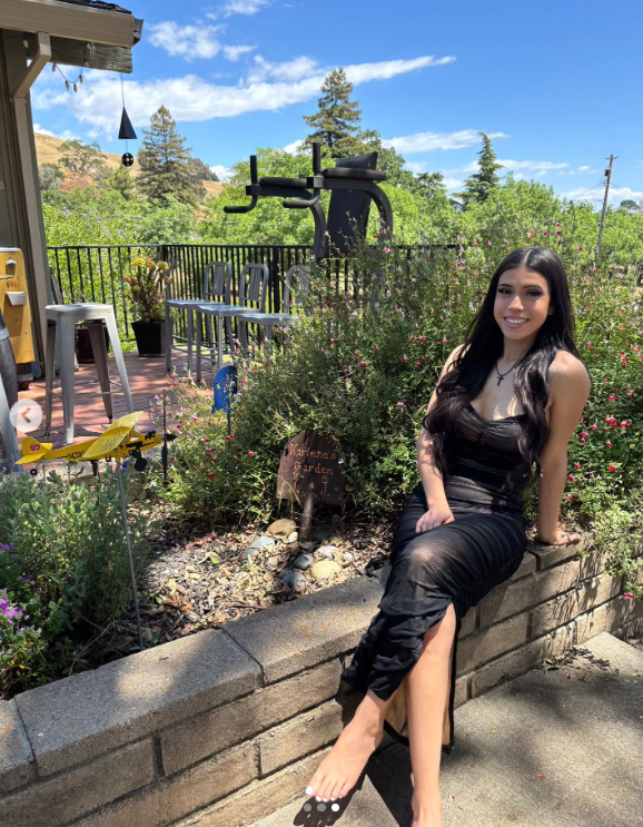 Marissa DiNapoli est assise dehors dans un jardin, souriante dans une robe noire près d'un panneau indiquant "Karlene's Garden", publié le 1er juillet 2025 | Source : Instagram/moniice__