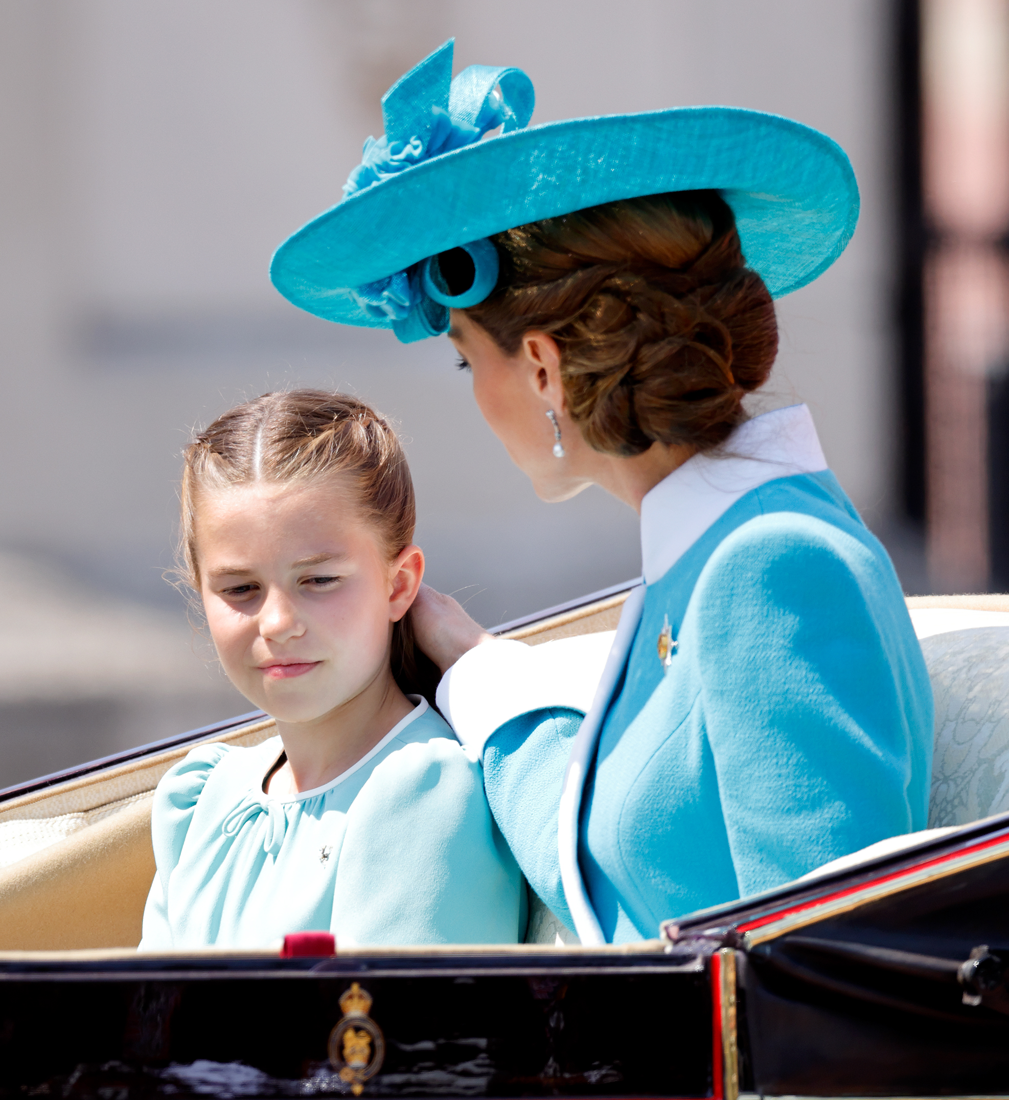 La princesse Charlotte de Galles et Catherine, princesse de Galles, retournent au palais de Buckingham. Catherine est radieuse dans sa robe couleur aquamarine, son chapeau à larges bords et ses cheveux bruns coiffés en une douce coiffure haute, tandis que Charlotte brille dans sa robe blanche et aquamarine et ses volumineux cheveux bruns, portés détachés avec une raie centrale soignée.