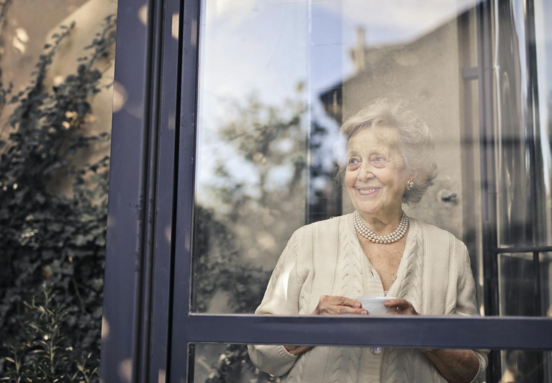 Une dame âgée souriante tenant sa tasse de thé et regardant par la fenêtre | Source : Pexels