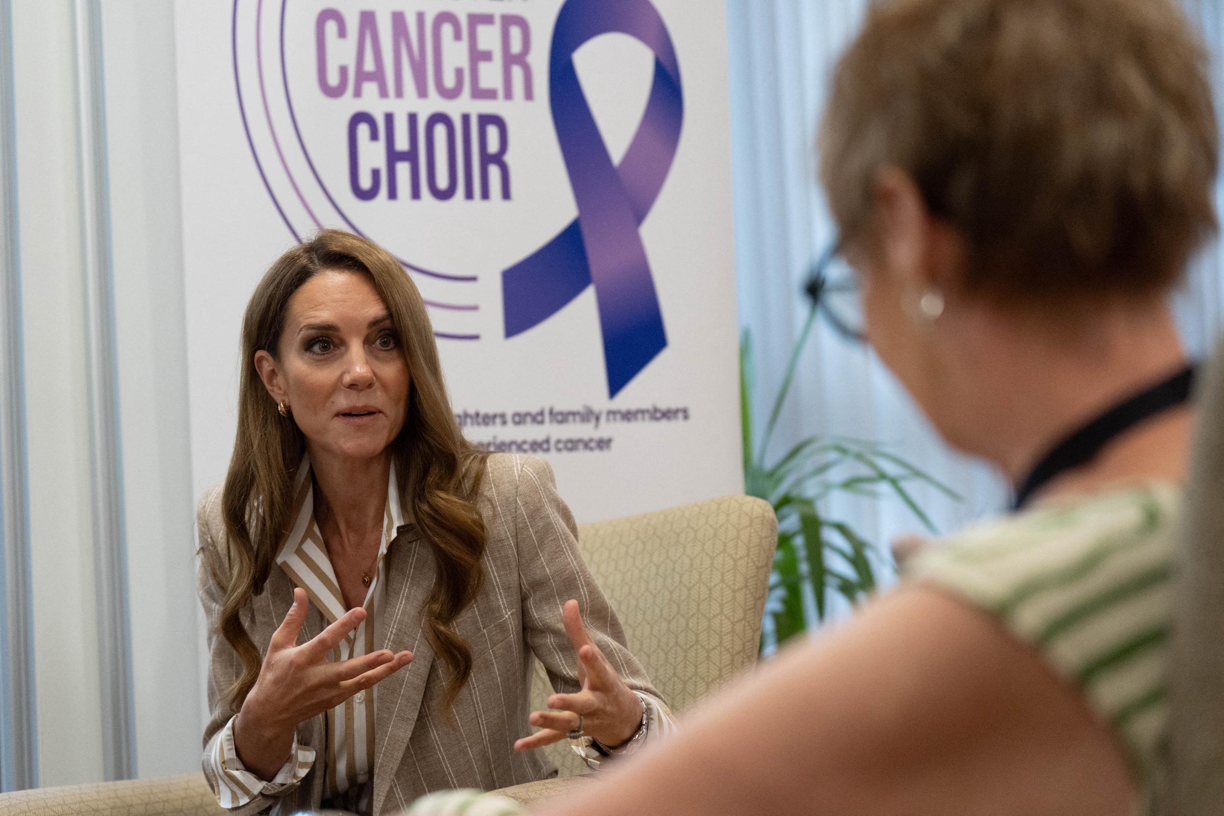 Assise à côté d'un panneau de sensibilisation au cancer, la princesse Catherine s'entretient avec une invitée. L'expression de la princesse de Galles est engageante et sincère.
