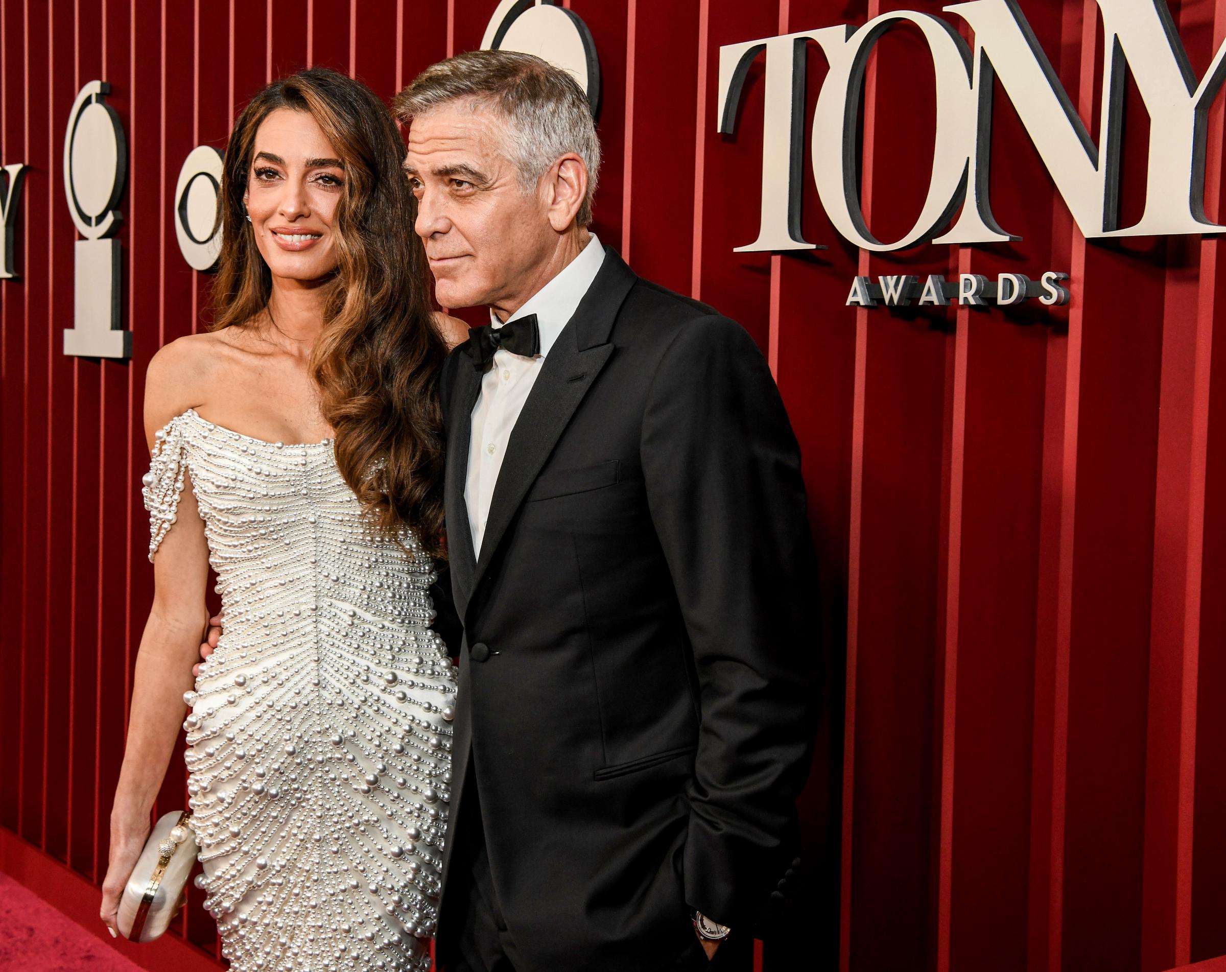 Amal Clooney offre un doux sourire, sa pochette sertie de perles à la main, tandis que George Clooney se tient près d'elle, les mains dans les poches, le regard perdu sur le côté.