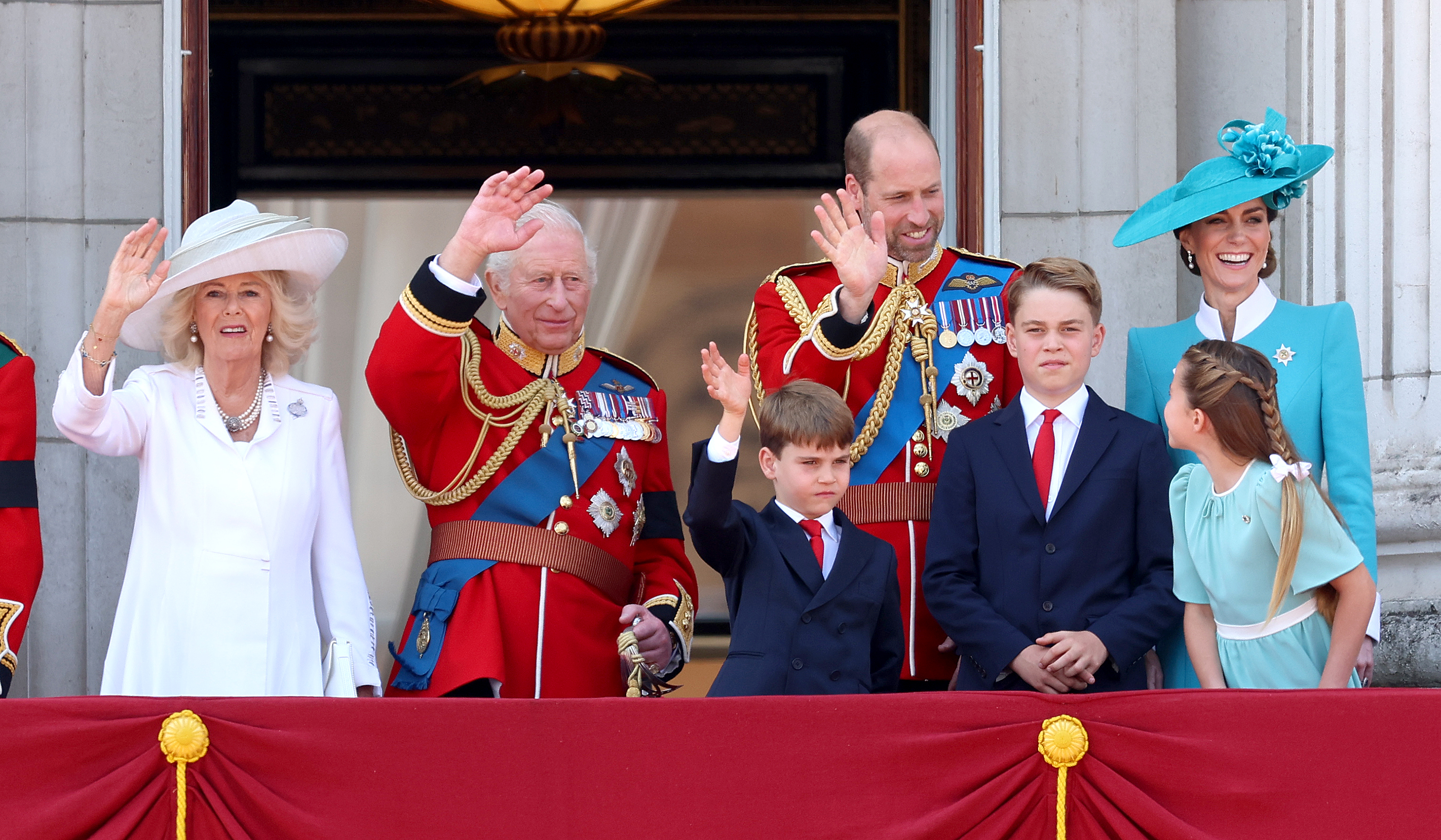 La reine Camilla, le roi Charles III, le prince Louis, le prince William, le prince George, la princesse Charlotte et la princesse Catherine saluent la foule lors du Trooping the Colour de 2025. La princesse Charlotte regarde son père, le prince George pose les poignets croisés, la princesse Catherine sourit et les autres membres de la famille royale saluent la foule.