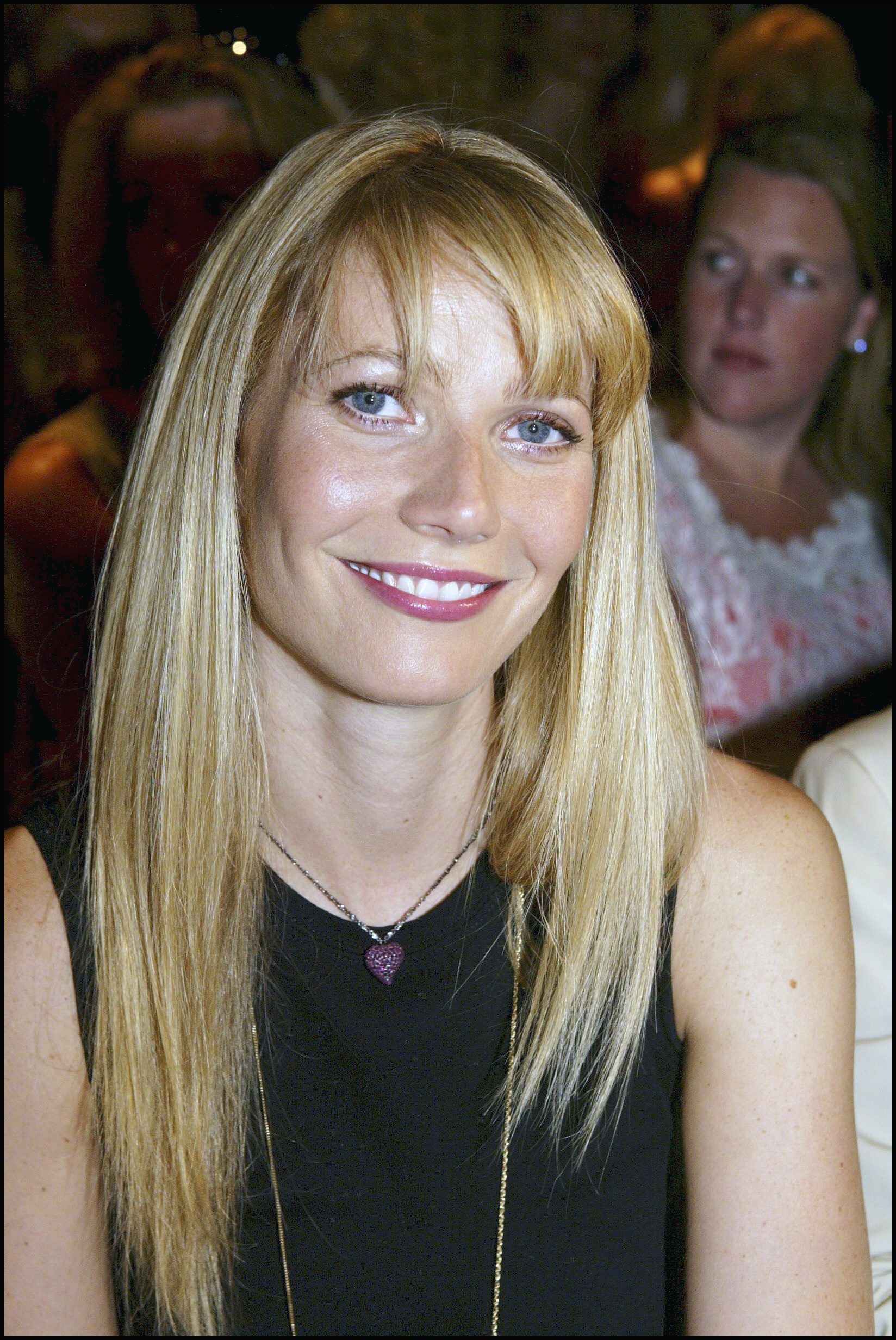 Gwyneth Paltrow assiste au défilé Valentino Haute Couture Automne-Hiver 2003-2004 en France, le 9 juillet 2003. Elle porte un haut noir sans manches associé à un délicat collier avec un pendentif en forme de cœur violet. Ses cheveux blonds raides sont coiffés avec une frange douce qui encadre son visage, tandis que son sourire radieux et ses lèvres brillantes couleur baie ajoutent une touche raffinée à son look.