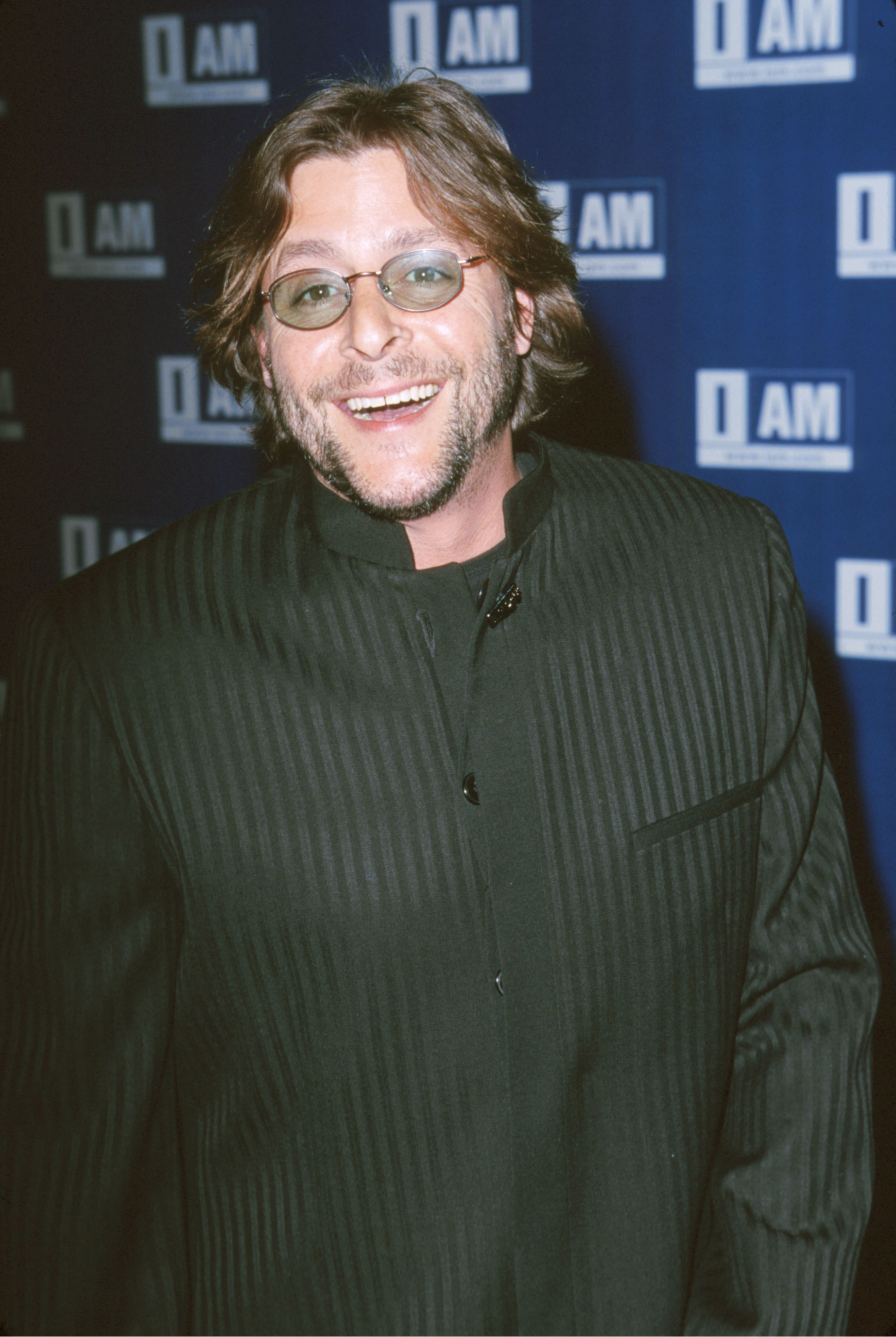 Avec ses lunettes ovales teintées, sa barbe de trois jours et sa veste à col mandarin à fines rayures foncées, l'acteur arborait un large sourire sur le tapis rouge lors de la soirée de lancement d'IAM.com à Hollywood, organisée aux Quixote Studios le 21 mars 2000. | Source : Getty Images