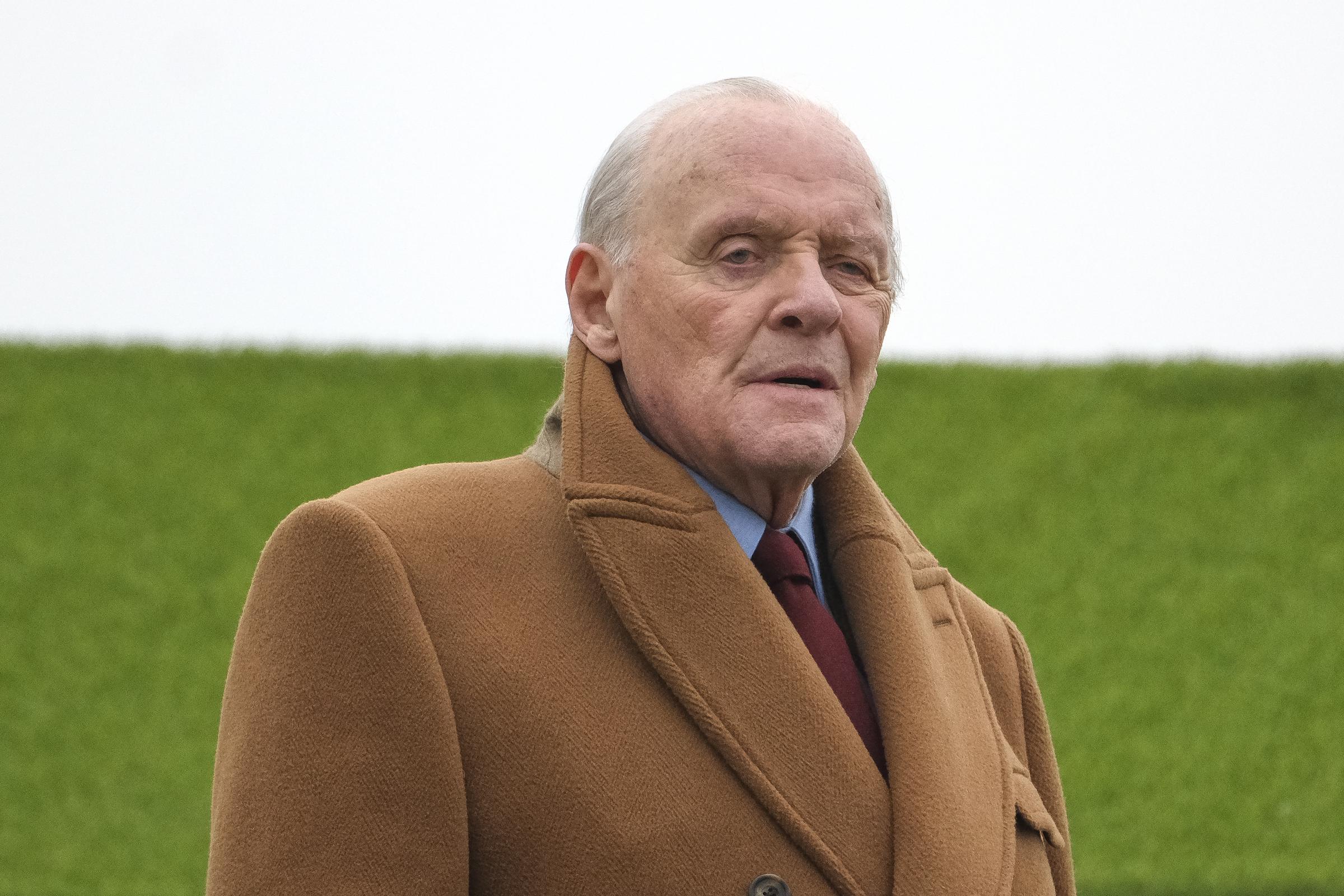 On voit Anthony Hopkins en train de tourner à l'hippodrome de Sandown Park, vêtu d'un riche manteau camel qui respire une élégance discrète. Son expression posée et son regard pensif capturent l'essence d'un homme profondément ancré dans son personnage. Sur fond de verdure éclatante, il se dresse comme une figure gracieuse et solennelle au milieu de la fraîcheur anglaise.