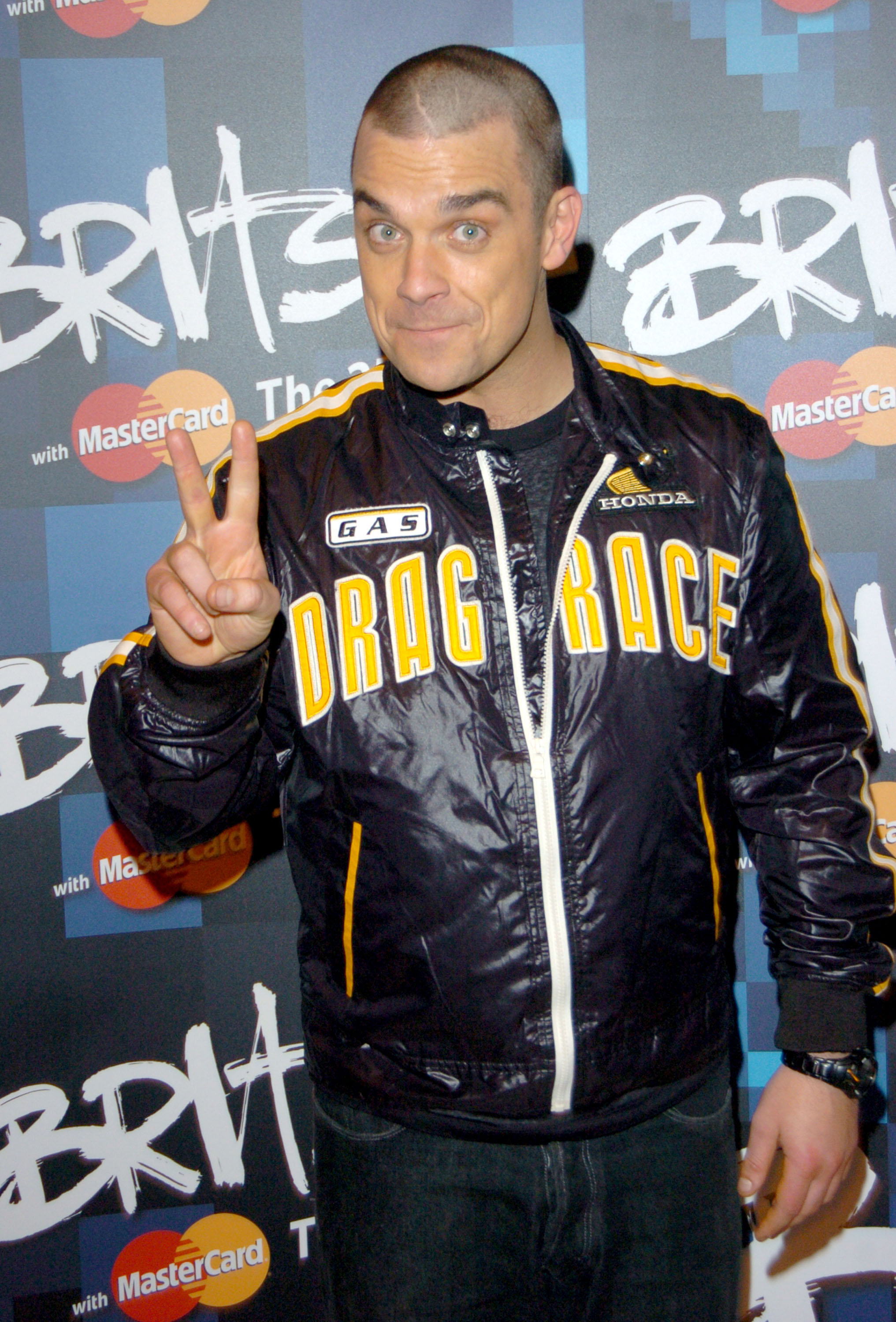 Robbie Williams pose aux Brit Awards 2005, faisant le signe de la paix dans une veste audacieuse « Drag Race ». Avec sa tête fraîchement rasée et son énergie espiègle, il prouve une fois de plus son talent pour allier espièglerie et style sur le tapis rouge.