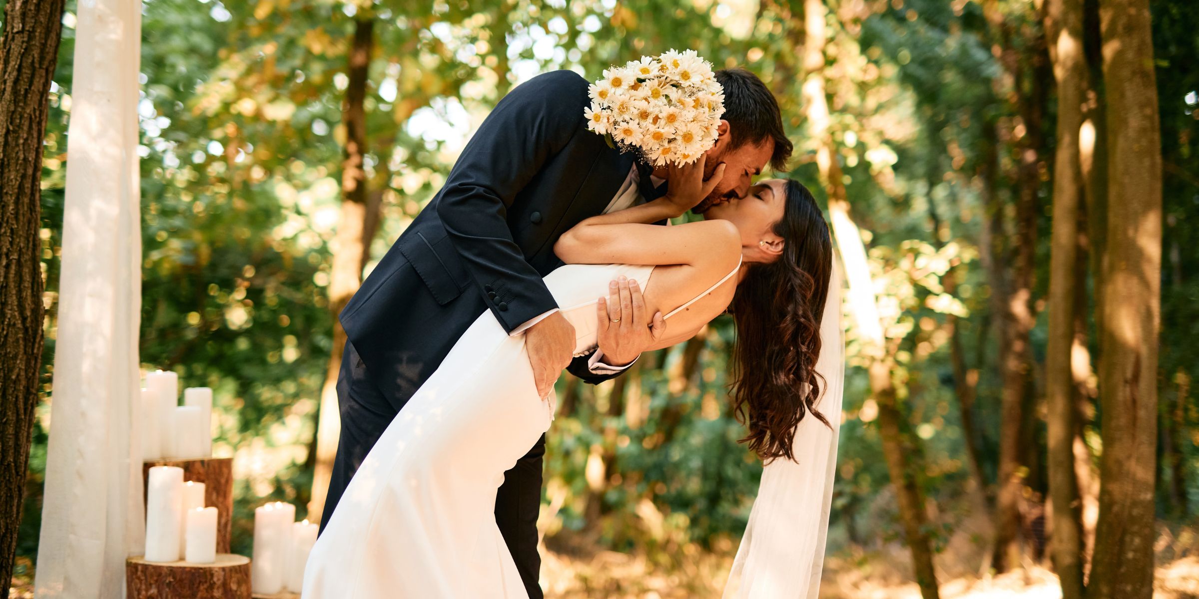Le marié, un bouquet à la main, penche la mariée vers lui pour l'embrasser lors d'une cérémonie de mariage romantique en forêt | Source : Getty Images