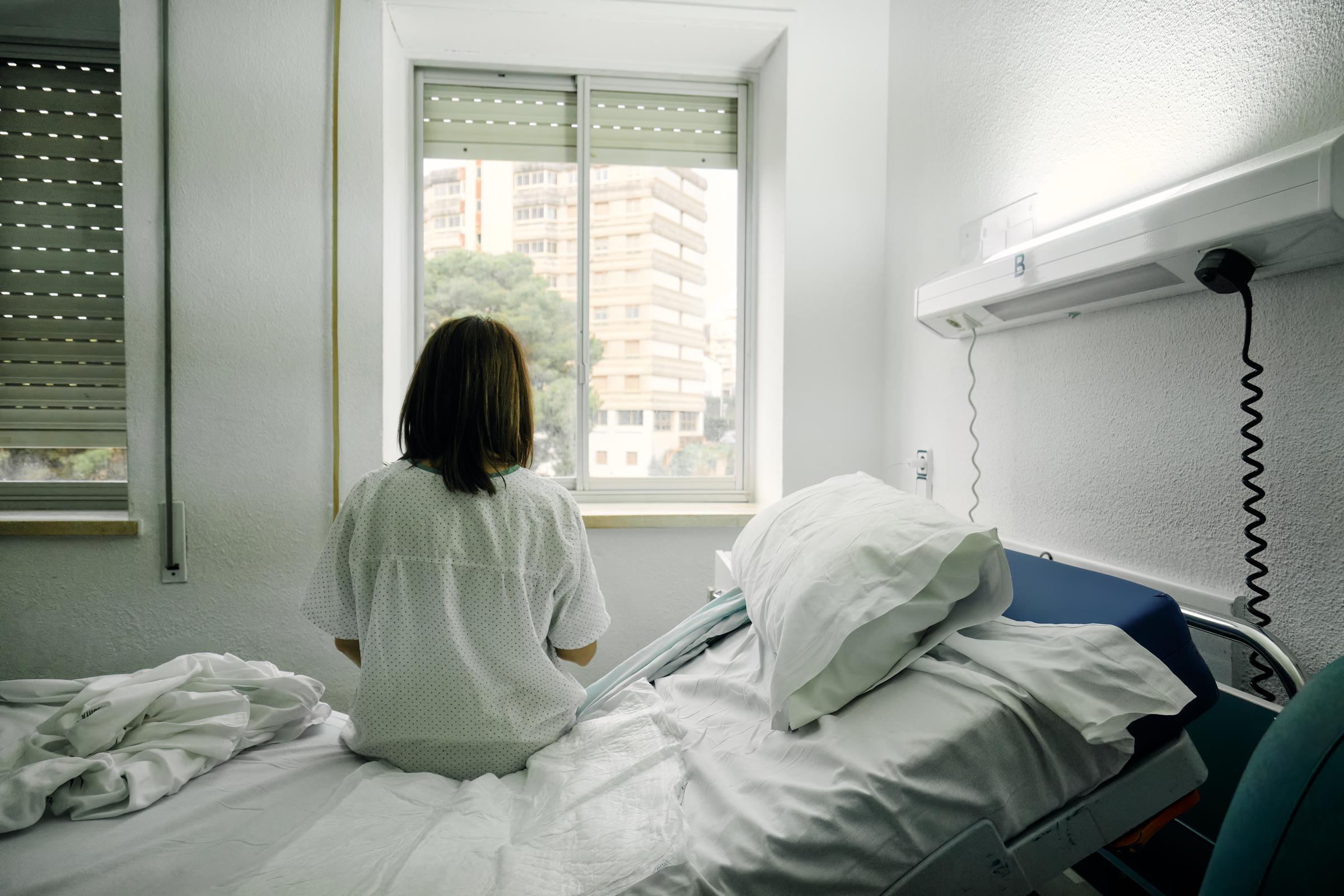 Une femme malade, seule, assise sur le lit d'une chambre d'hôpital, regardant par la fenêtre I Photo d'illustration I Source : Getty Images