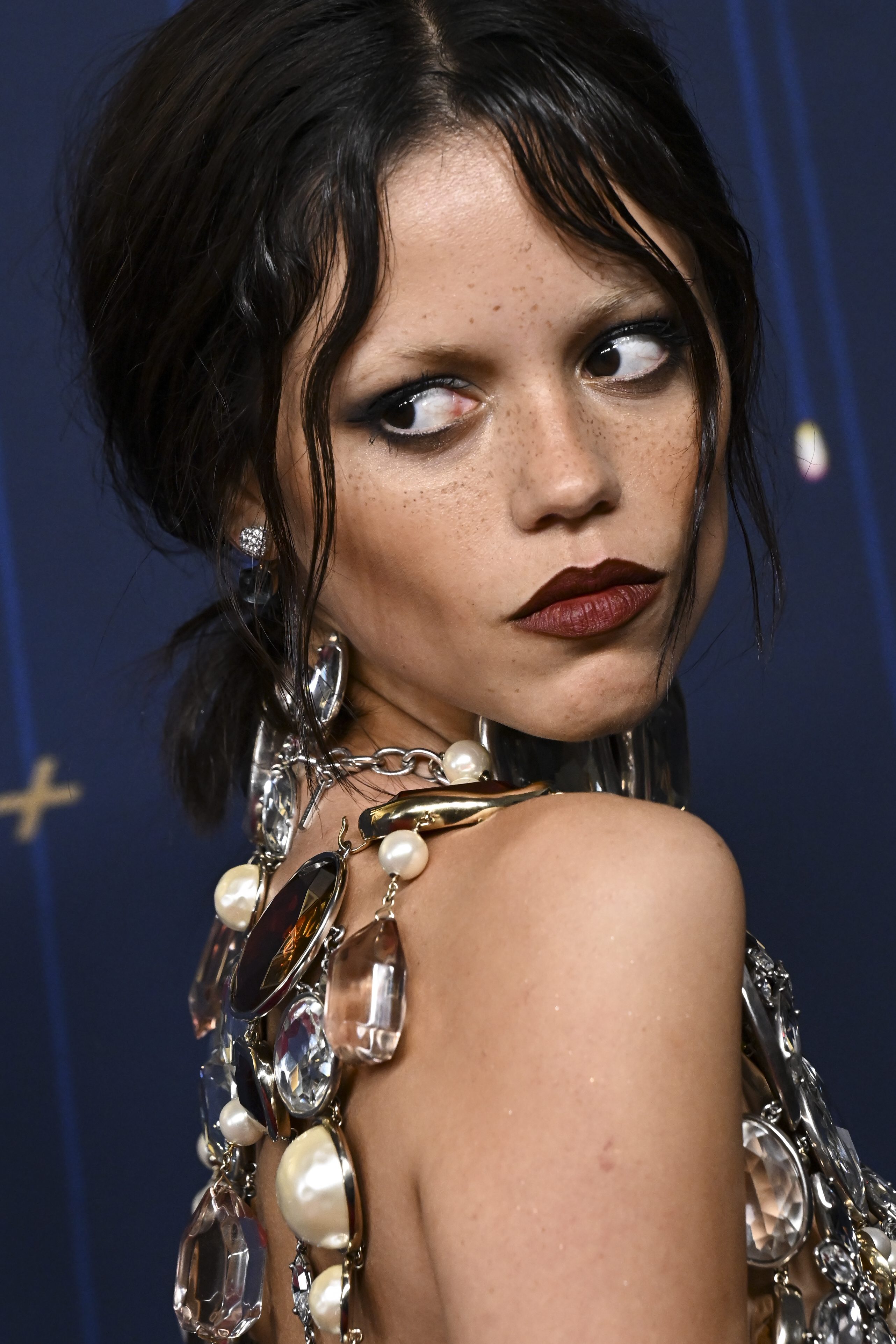 Le visage de Jenna Ortega photographié lors de la 77e cérémonie des Primetime Emmy Awards à Los Angeles, en Californie. Elle porte un rouge à lèvres bordeaux foncé et ses cheveux noirs semblent ébouriffés et séparés par une raie au milieu.