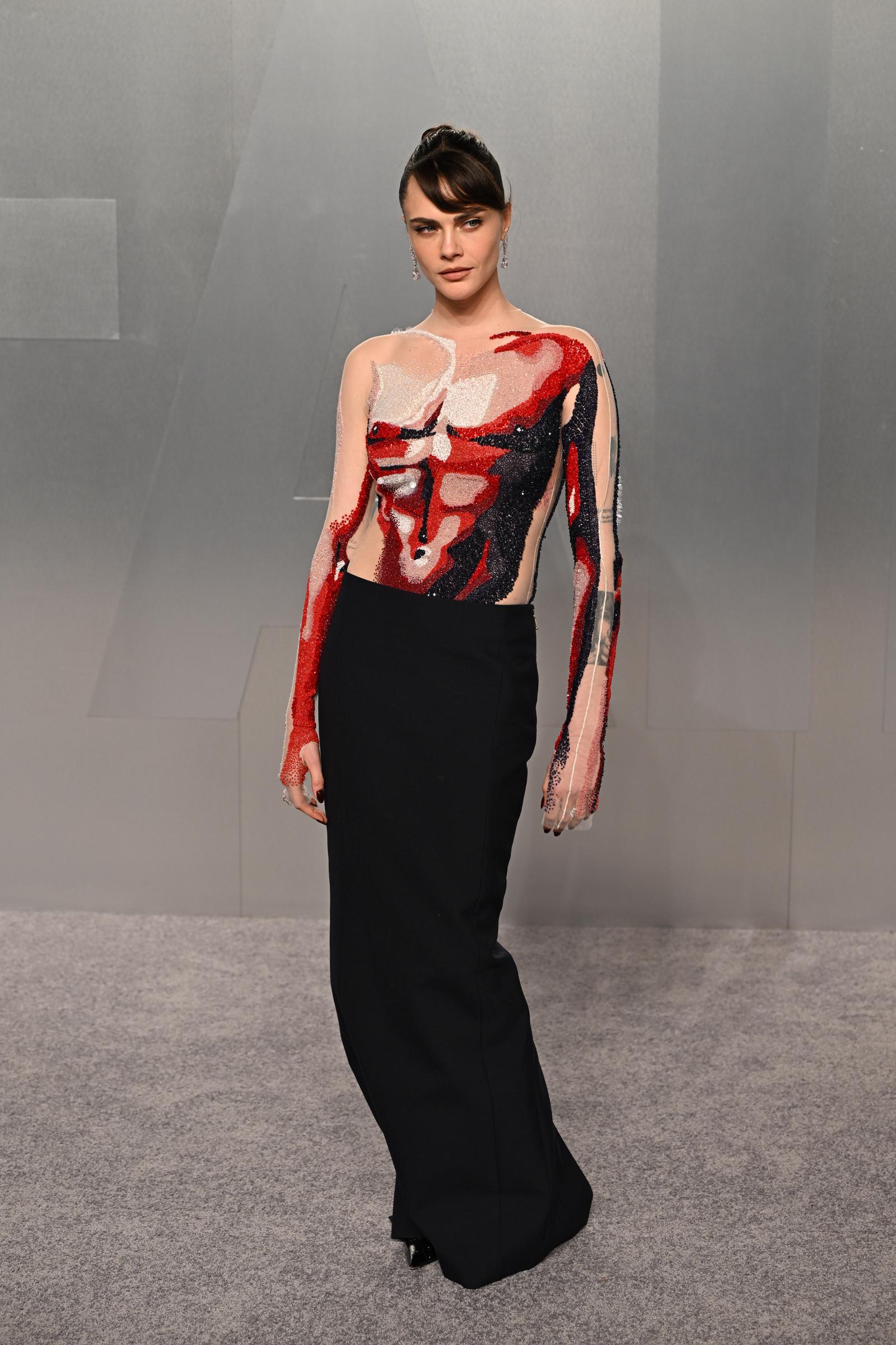 Cara Delevingne pose lors de la soirée Vanity Fair des Oscars, vêtue d'un body nude transparent à manches longues, orné de motifs picturaux abstraits en perles et paillettes rouges, blanches et noires sur le torse et les bras, qu'elle a associé à une jupe droite noire simple tombant jusqu'au sol et à de longues boucles d'oreilles en forme de diamant.