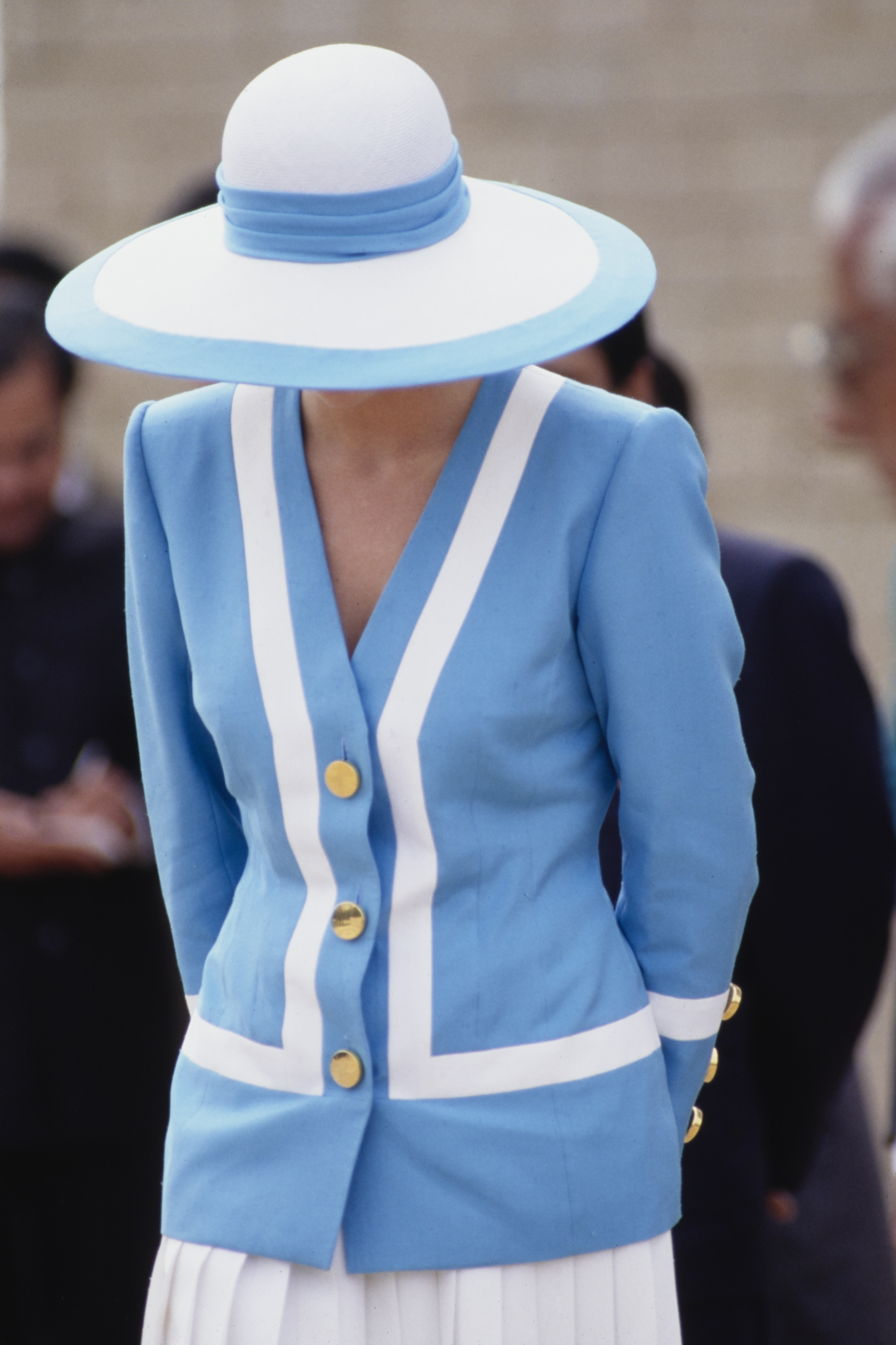 La princesse Diana se tient debout avec un calme tranquille au cimetière militaire de Delhi, vêtue de son tailleur bleu et blanc caractéristique et d'un chapeau à larges bords. L'image capture sa grâce solennelle au milieu d'un cadre tranquille, reflet d'un souvenir respectueux.
