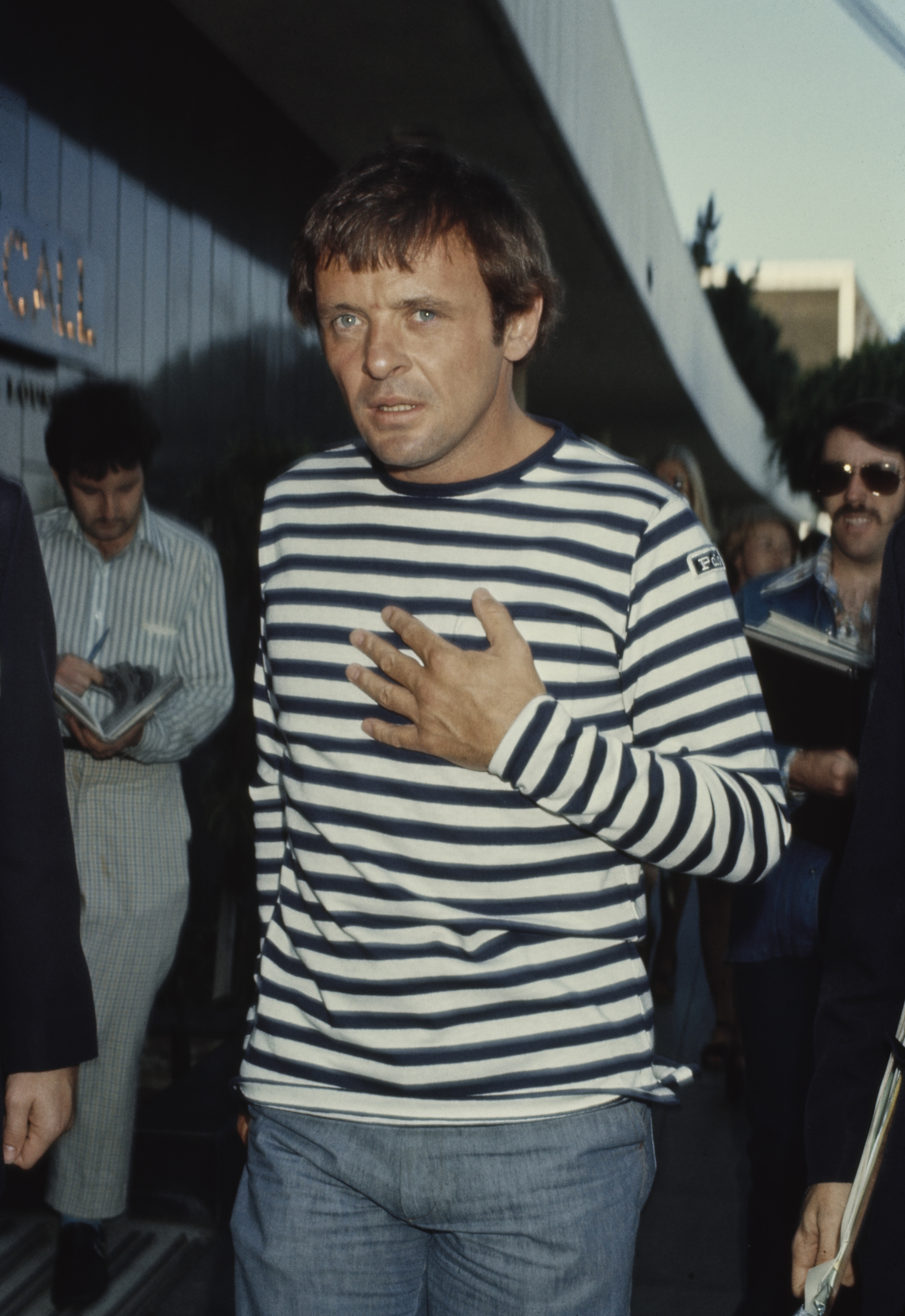 Anthony Hopkins avance d'un pas décidé, le regard partagé entre réflexion et intensité. Vêtu d'un t-shirt décontracté à rayures bleu marine et blanches, il dégage un charme naturel teinté d'une touche de mystère. La lumière déclinante et les spectateurs qui l'entourent confèrent à la scène une atmosphère cinématographique empreinte d'une attente silencieuse.