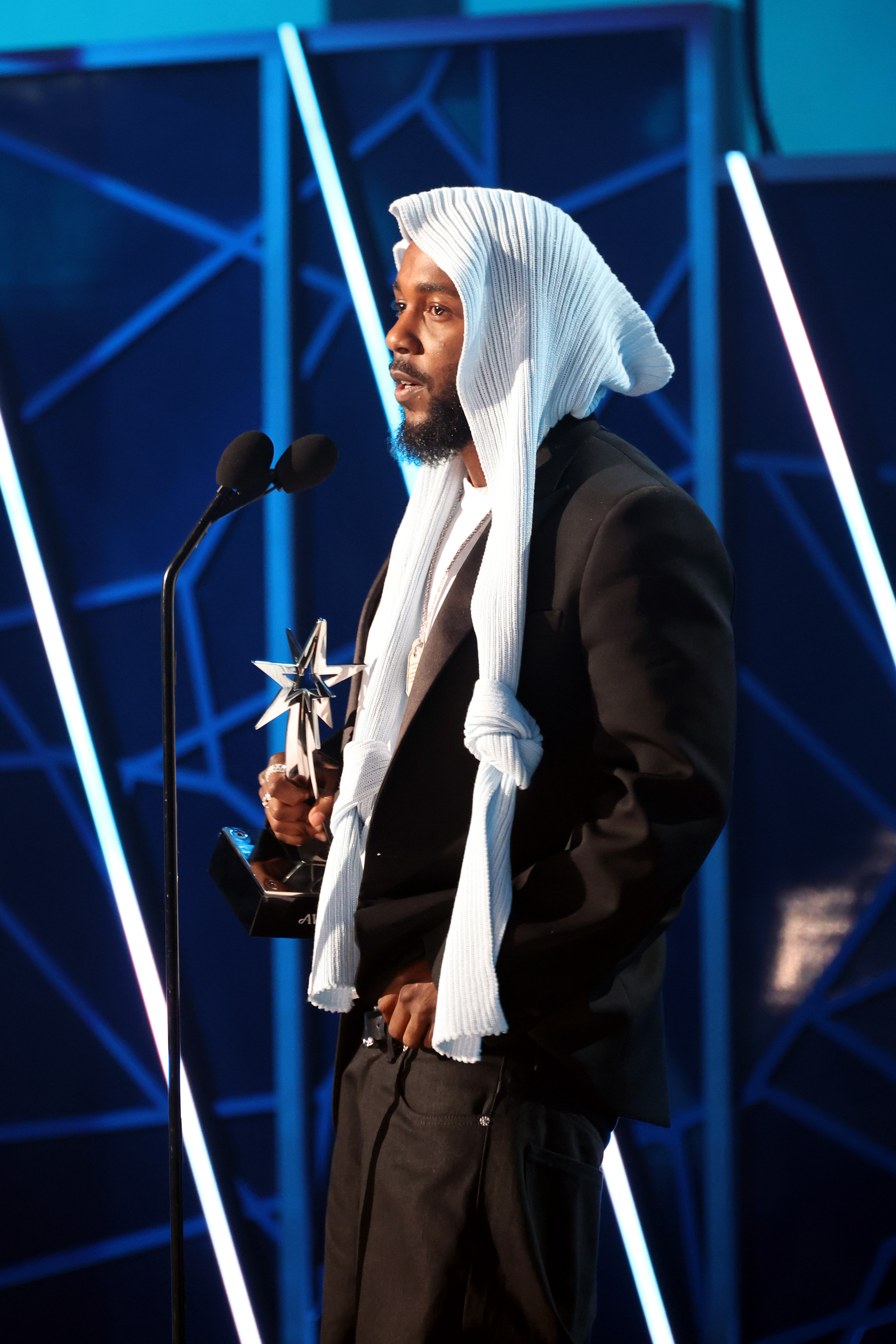 Avec une présence à la fois calme et puissante, Kendrick Lamar s'adresse au public après avoir remporté la victoire aux BET Awards 2025. Vêtu d'un ensemble noir et blanc, l'artiste tenait fièrement son trophée devant la toile de fond géométrique rougeoyante.