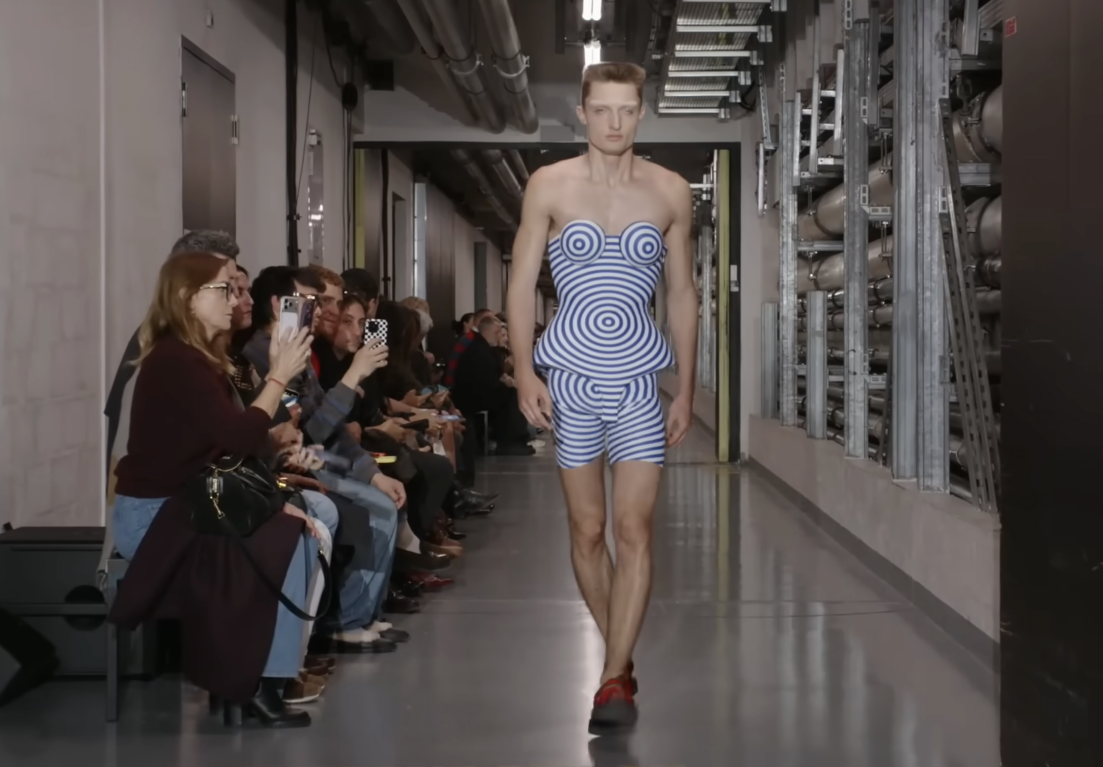 Un mannequin présente un autre modèle futuriste qui semble avoir été imprimé en 3D. Le haut en forme de péplum est associé à un short assorti, créant un look audacieux et moderne sur le mannequin masculin. Extrait d'une publication datée du 5 octobre 2025. Source : Youtube/@jeanpaulgaultier