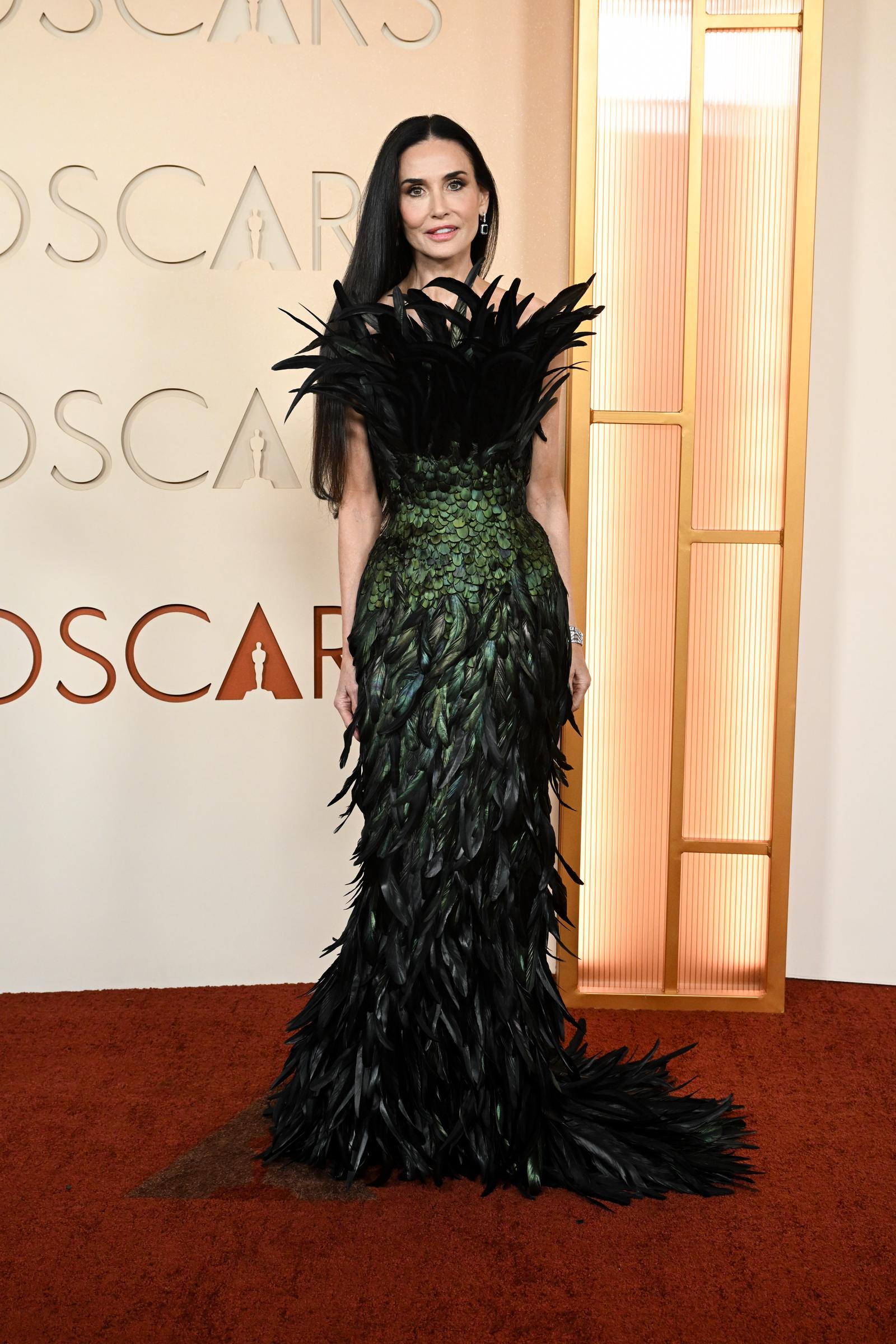 Demi Moore arrive à la 98e cérémonie des Oscars vêtue d'une robe longue vert foncé entièrement composée de plumes irisées, avec un col à plumes spectaculaire et surdimensionné qui s'épanouit au niveau des épaules et une traîne à plumes qui s'étend derrière elle.