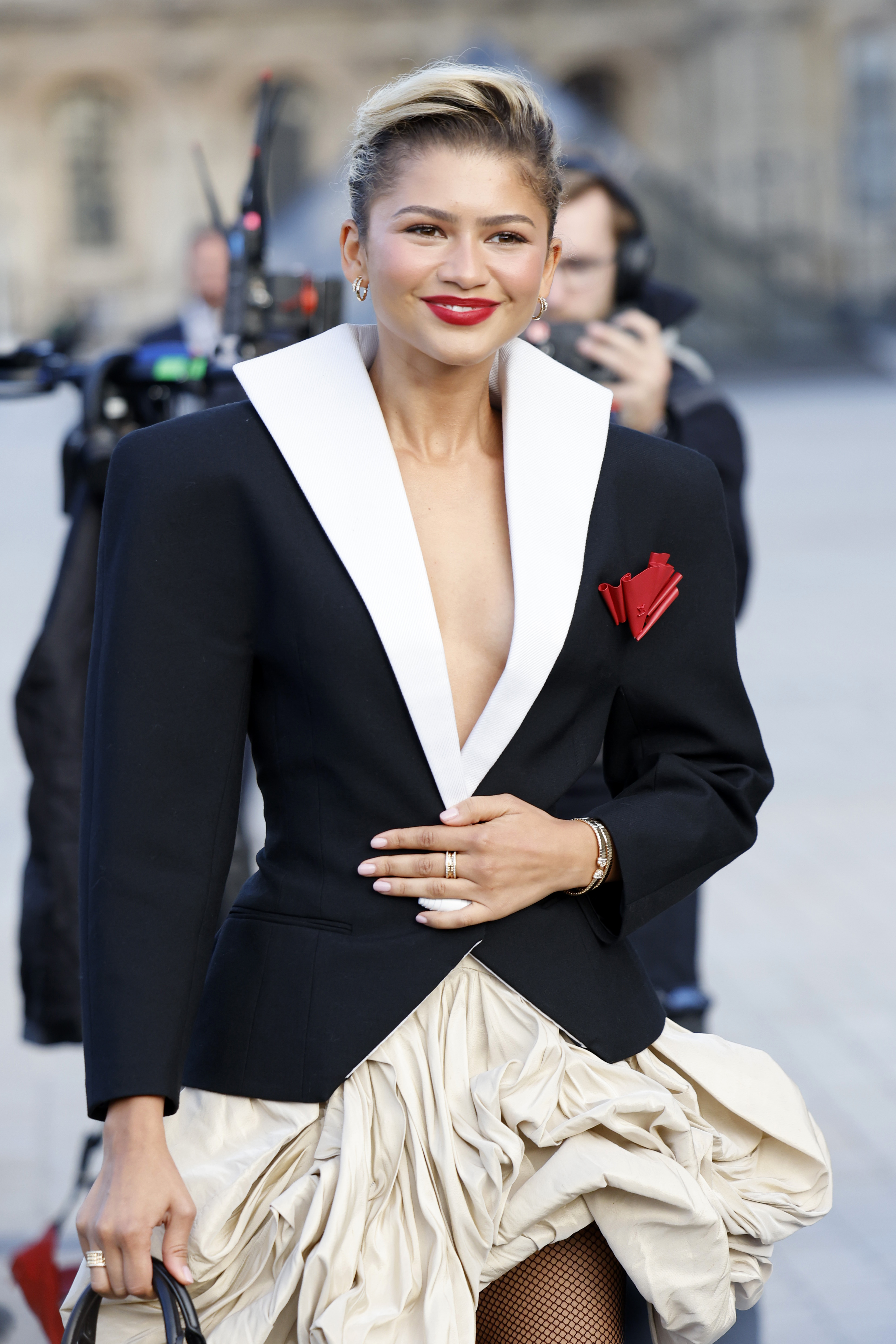 Zendaya assiste au défilé femme printemps/été 2025 de la Fashion Week de Paris, en France, vêtue d'un blazer noir sculpté à l'encolure plongeante, aux épaules exagérées et au col blanc montant. Le blazer, fermé par un seul bouton cylindrique blanc, est orné d'une application rouge en forme de rose. Elle le porte sur une jupe bouffante beige.