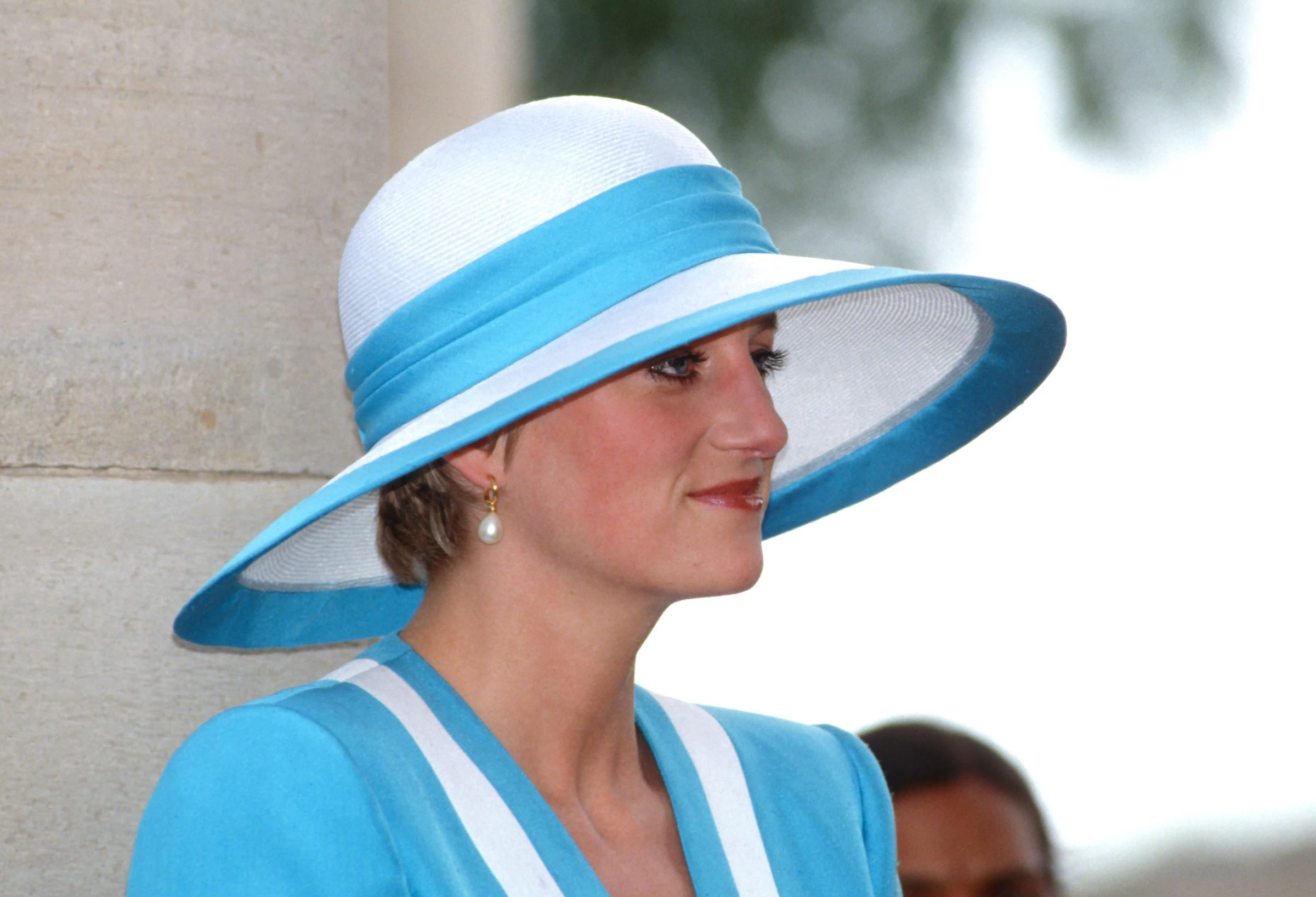 Dans un gros plan pris lors de sa visite au cimetière militaire de Delhi en 1992, l'expression sereine de la princesse Diana occupe le devant de la scène. Vêtue d'un élégant tailleur bleu et blanc signé Catherine Walker et coiffée d'un chapeau sculpté Philip Somerville, la caméra capture la dignité tranquille de son visage, élégamment mis en valeur par son style caractéristique.