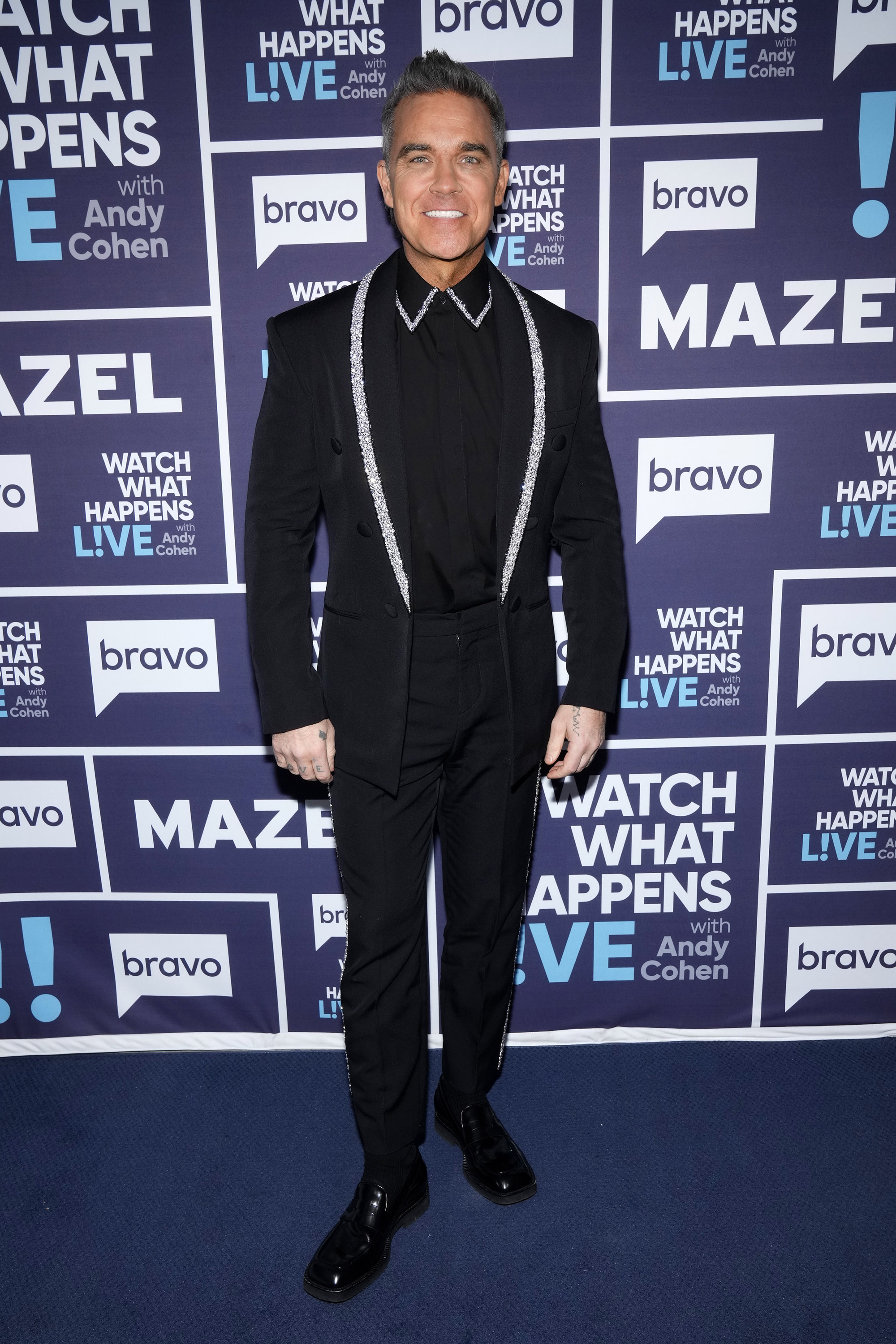 Robbie Williams participe à l'émission « Watch What Happens Live With Andy Cohen » en 2025, vêtu d'un élégant costume noir aux finitions scintillantes. Souriant devant les journalistes, il allie un style raffiné à son charisme habituel.