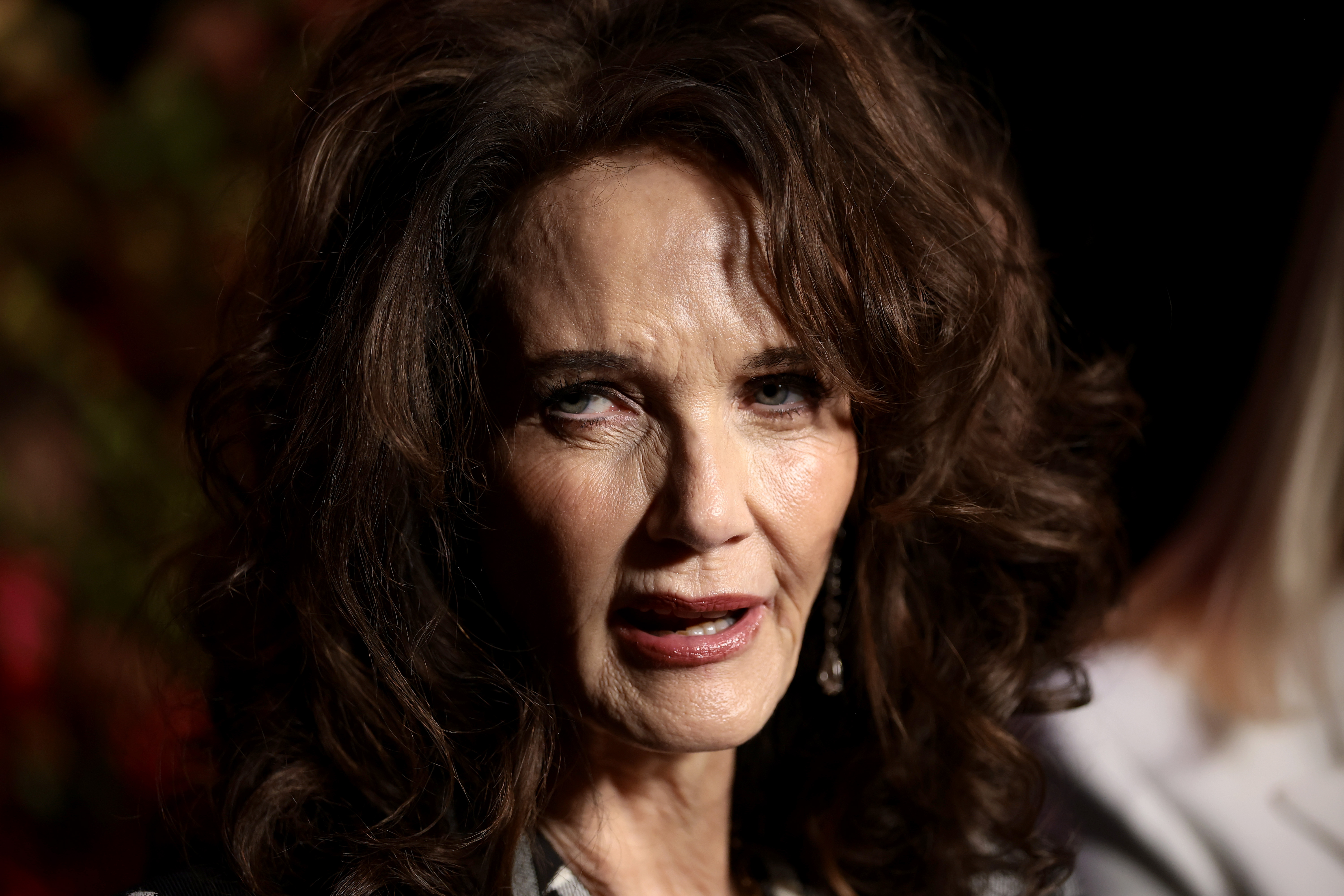 Lynda Carter est photographiée de près, l'air posé et contemplatif sous un éclairage doux et tamisé. Ses boucles volumineuses encadrent son visage, tandis que son maquillage subtil et ses boucles d'oreilles pendantes complètent son look raffiné et discret.