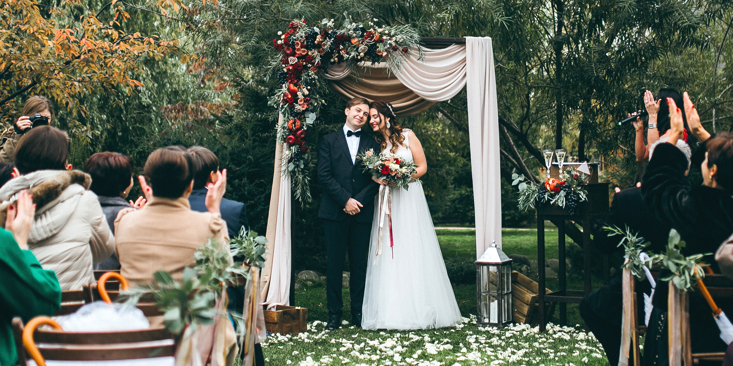 Une mariée et un marié se tenant sous une arche florale et souriant tandis que les invités applaudissent pendant leur cérémonie de mariage en plein air | Source : Shutterstock