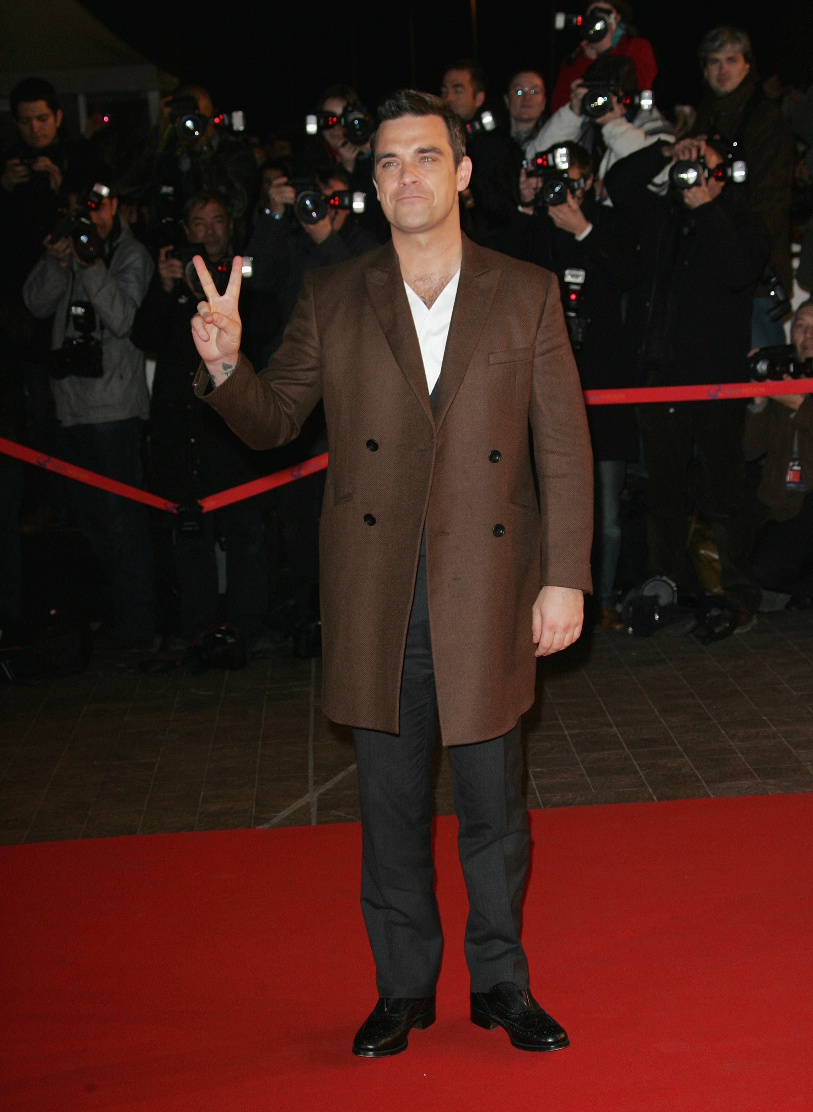 Robbie Williams aux NRJ Music Awards à Cannes, en France, le 23 janvier 2010. Arborant un signe de paix sur le tapis rouge dans un élégant manteau marron et une chemise blanche impeccable, il avait tout à fait l'air d'une star chevronnée sous les feux de la rampe.