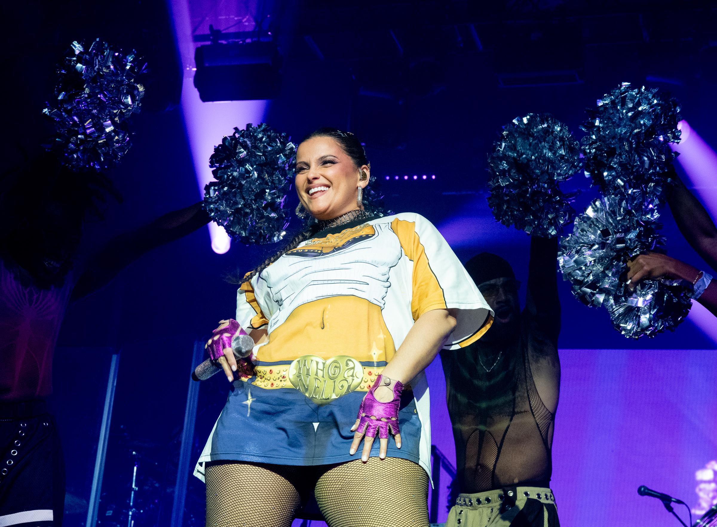 Furtado est accompagnée de danseurs tenant des pompons tandis qu'elle se produit vêtue d'un t-shirt oversize imprimé de dessins animés et de collants résille. Elle sourit tout en tenant un micro, portant des gants rose vif et une ceinture au design audacieux « Whoa Nelly » sur sa tenue.