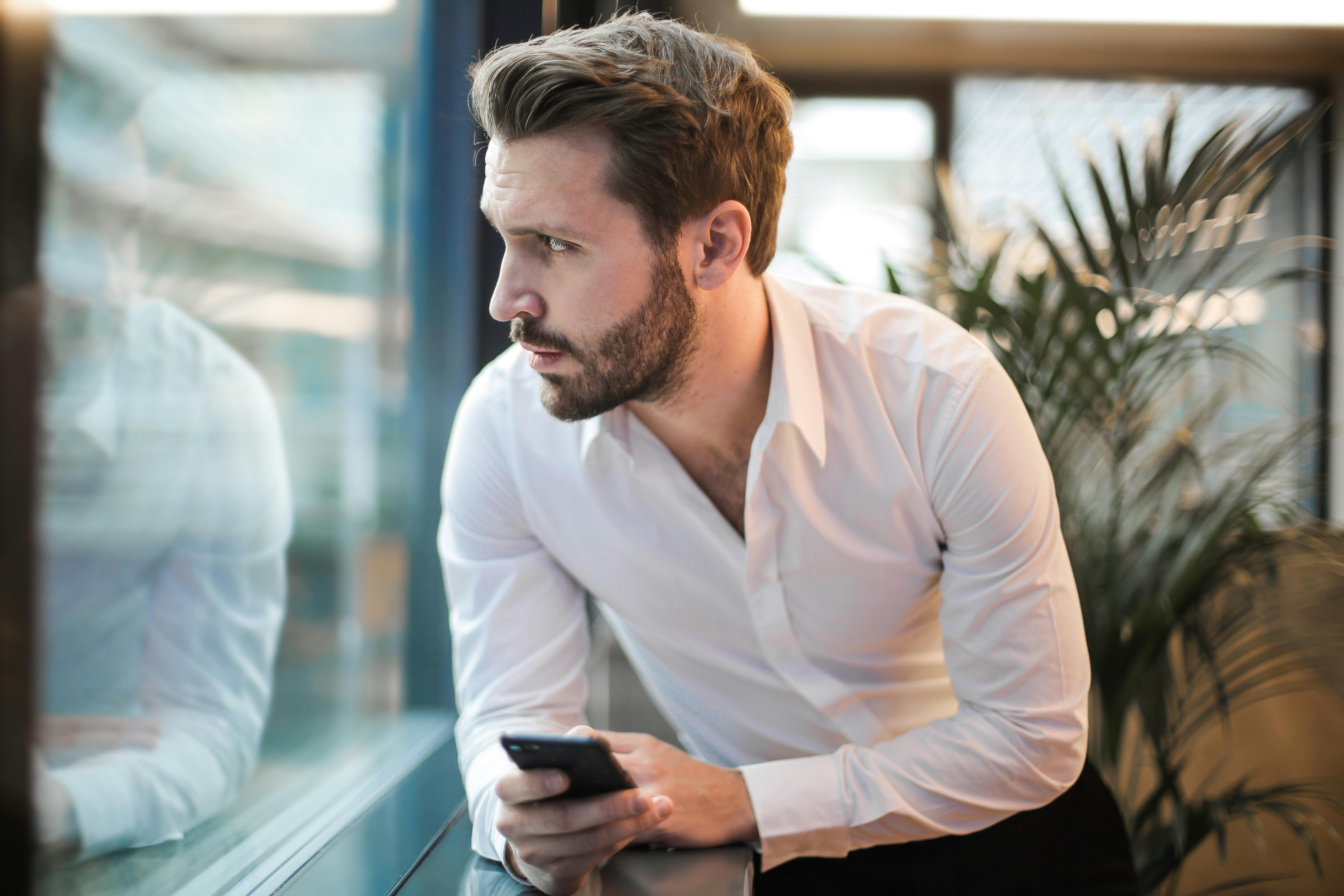 Un homme qui regarde par la fenêtre, le téléphone à la main | Source : Pexels