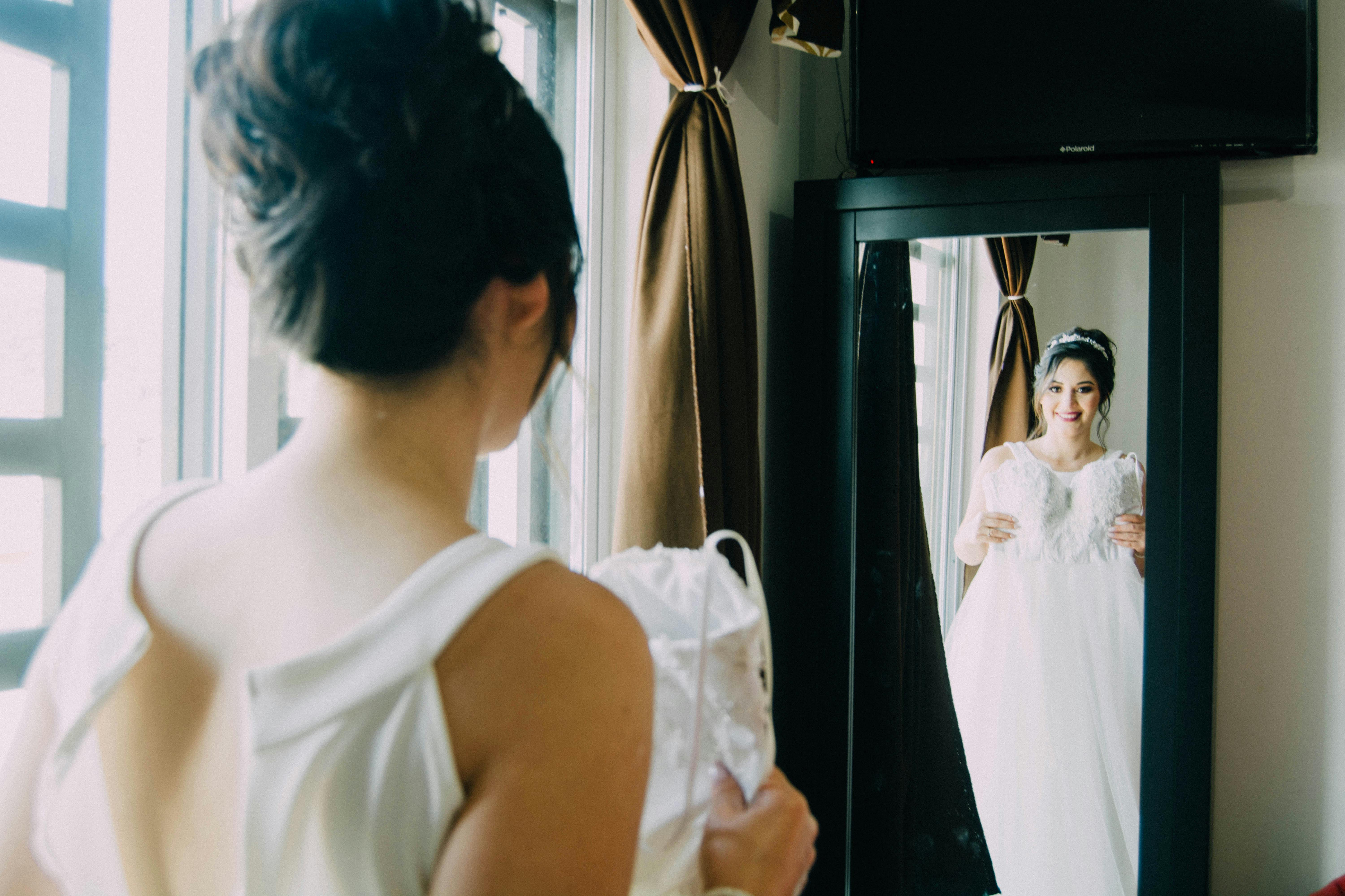 Une mariée heureuse tenant sa robe de mariée tout en se regardant dans un miroir | Source : Pexels